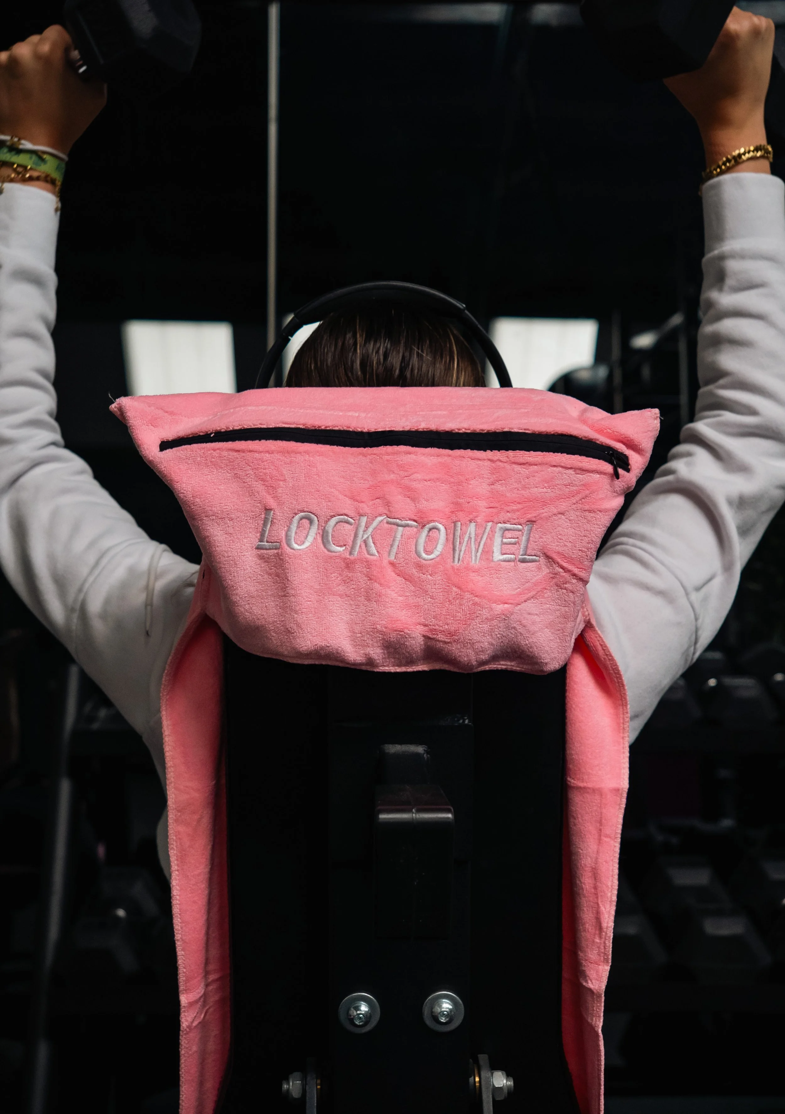 Persoon in sportschool met roze handdoek met de tekst 'LOCKTOWEL' op de rug, met armen omhoog die gewichten vasthouden.