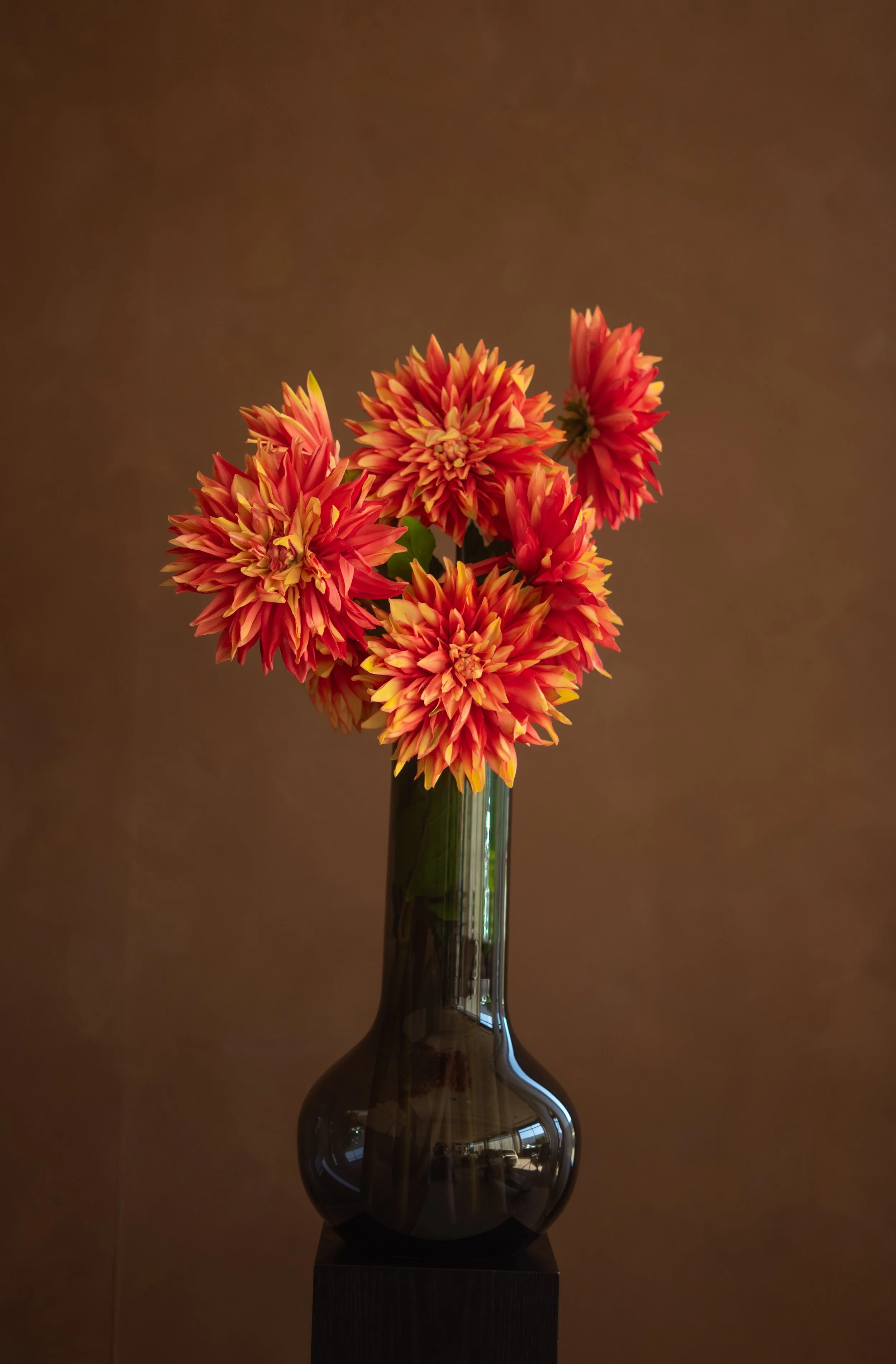 Oranje en gele bloemen in een groene glazen vaas op een zwarte standaard, tegen een bruin achtergrond. Floweris