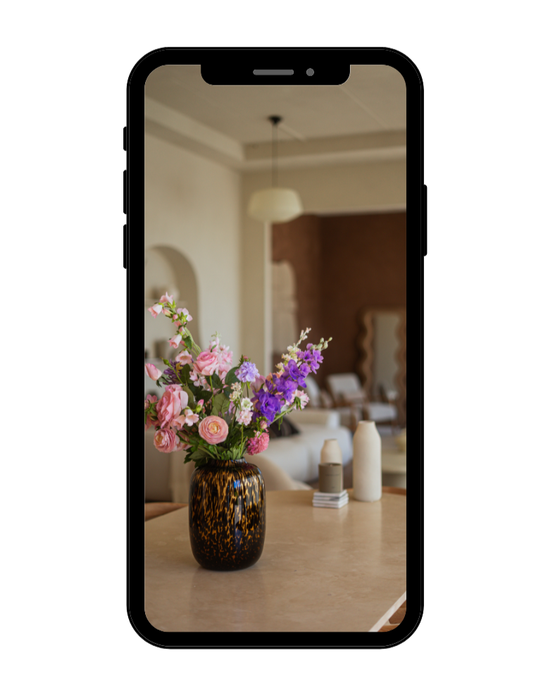 Een smartphone toont een foto van een bloemenvase met roze en paarse bloemen op een tafel in een interieur met een witte plafondlamp en een spiegel op de achtergrond.