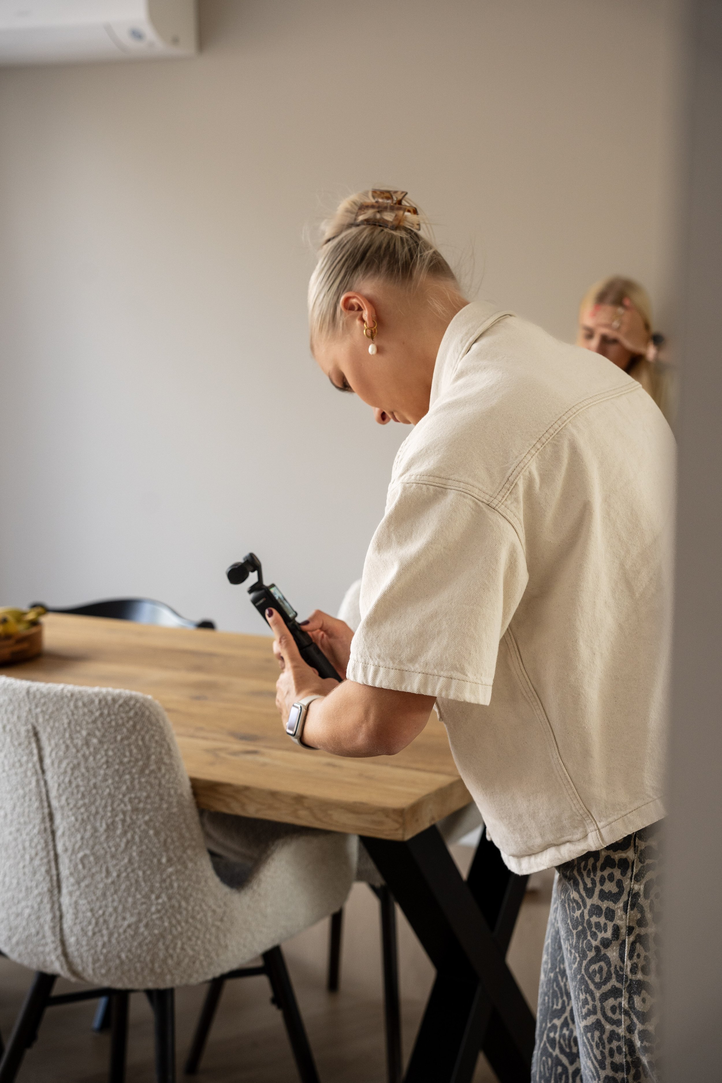 Een vrouw met een blonde paardenstaart, gouden oorbellen en een smartwatch leest op haar telefoon in een moderne eetkamer, terwijl een andere vrouw op de achtergrond zichtbaar is.