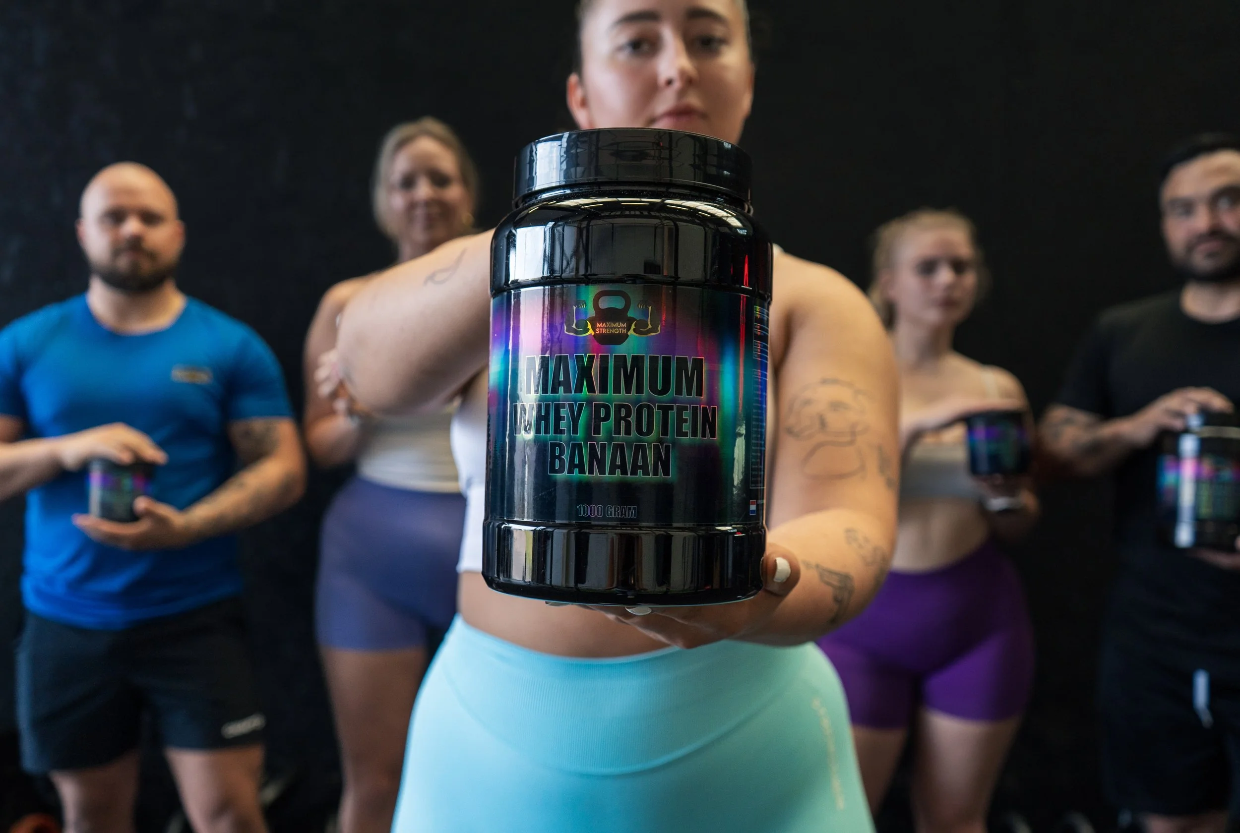 Vrouw houdt een pot van dieetvoeding met label 'Maximum Whey Protein Banaan' voor de camera, met vier mensen op de achtergrond in een sportschool. Sportsupplementen.