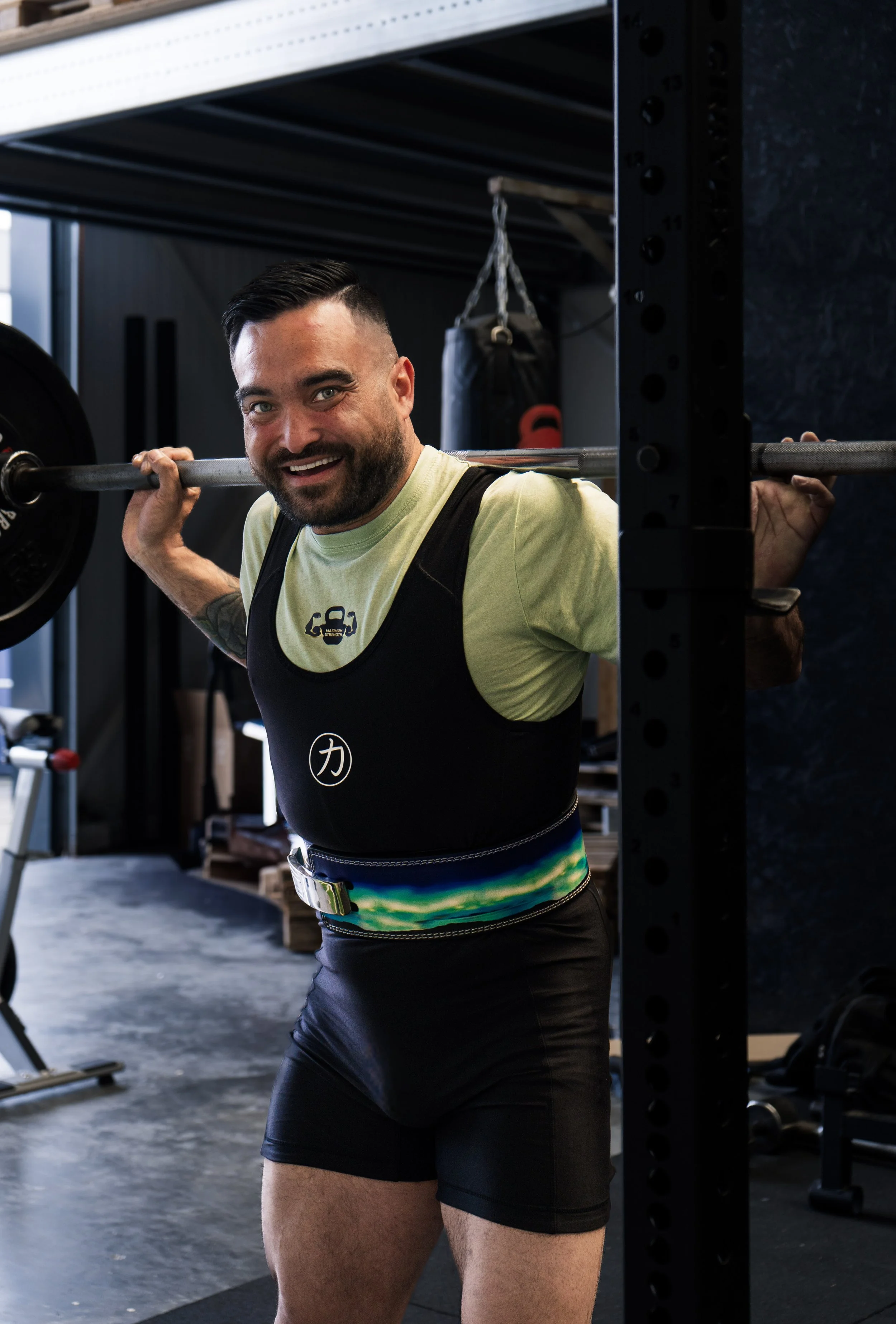 Man met sporten in sportschool, hij staat in een squatpositie met een halter op zijn schouders, glimlachend.