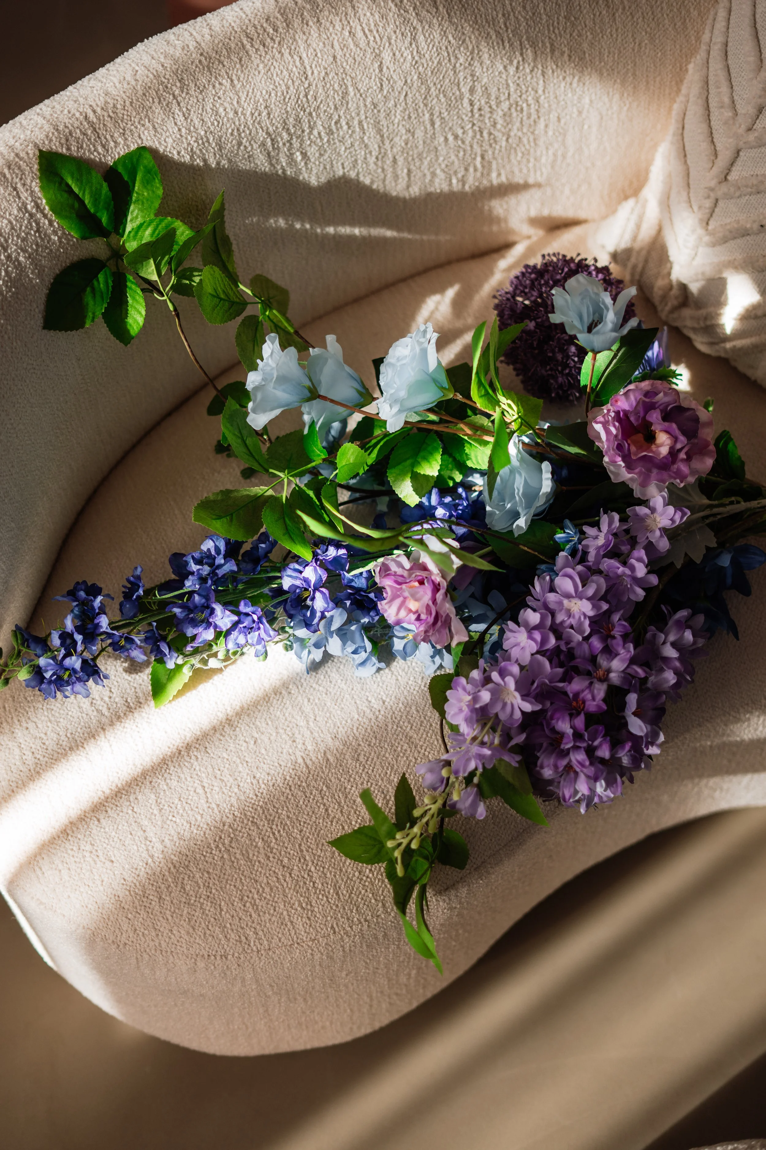 Een bos bloemen met blauwe, paarse, witte en roze bloemen en groene bladeren ligt op een beige bank met een zachte textuur, met zonlicht dat zachte schaduwen werpt. Floweris