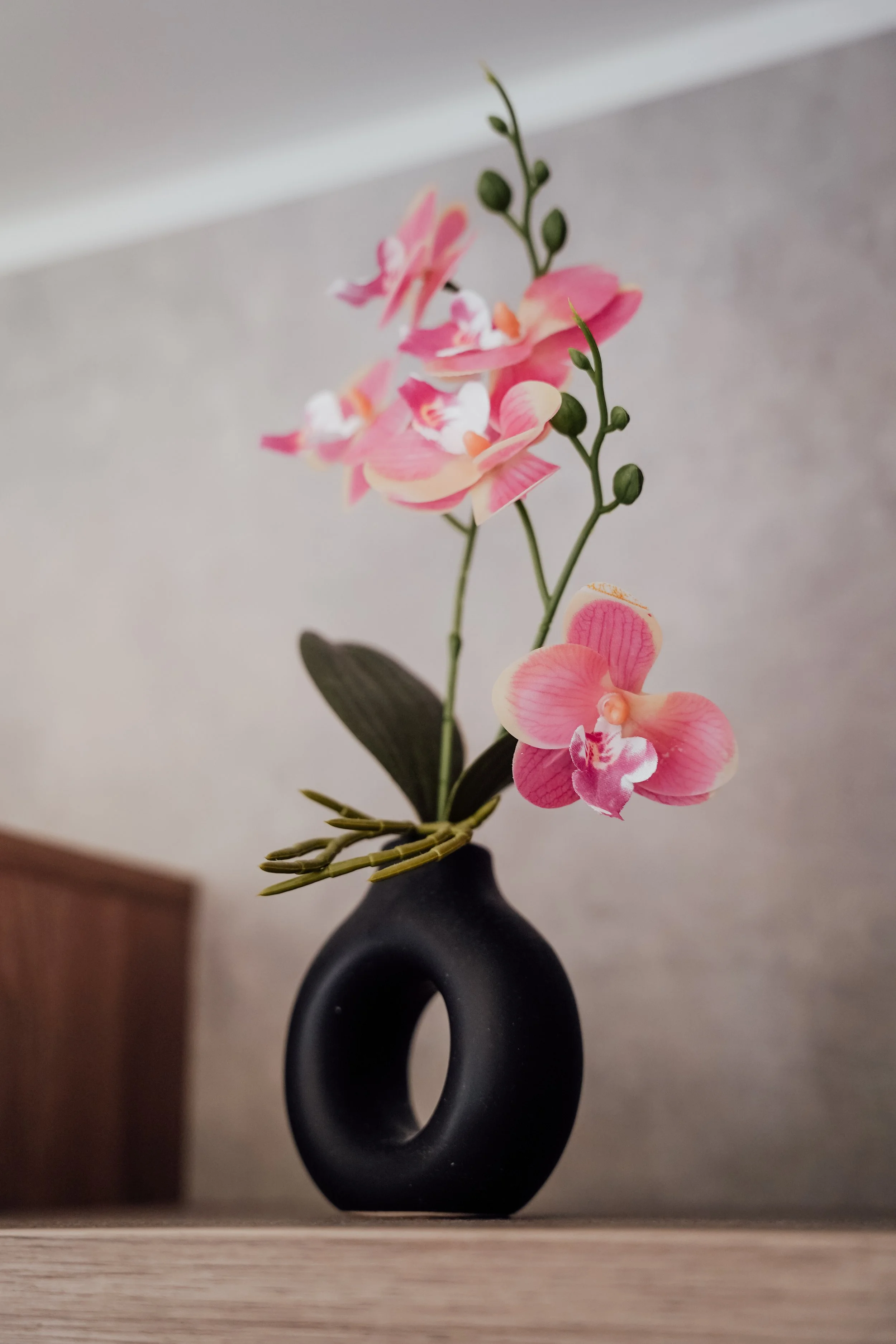 Een bloemstuk met roze orchideeën in een zwart, ronde vaas op een houten oppervlak, met een neutrale achtergrond. Vakantiepark. Chalet.