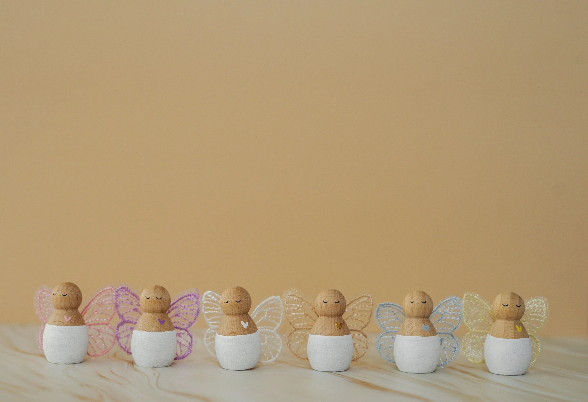 Vijf houten engel figuren met gekleurde vleugels en witte rokken, elk met een klein hartje op de borst, uitgelijnd op een licht houten oppervlak tegen een beige achtergrond.