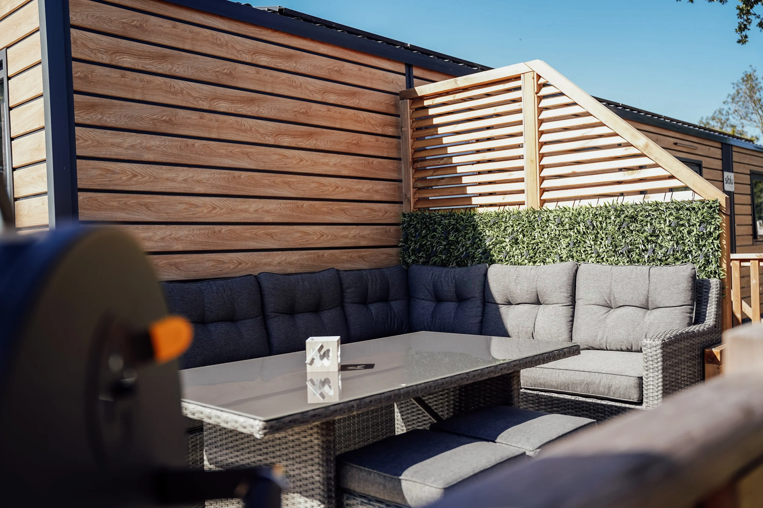 Een buitenzitje met een L-vormige bank met kussens, een tafel met een glazen blad, en een houten schutting met groen gewas ertegen, onder een blauwe hemel. Vakantiepark. Chalet.