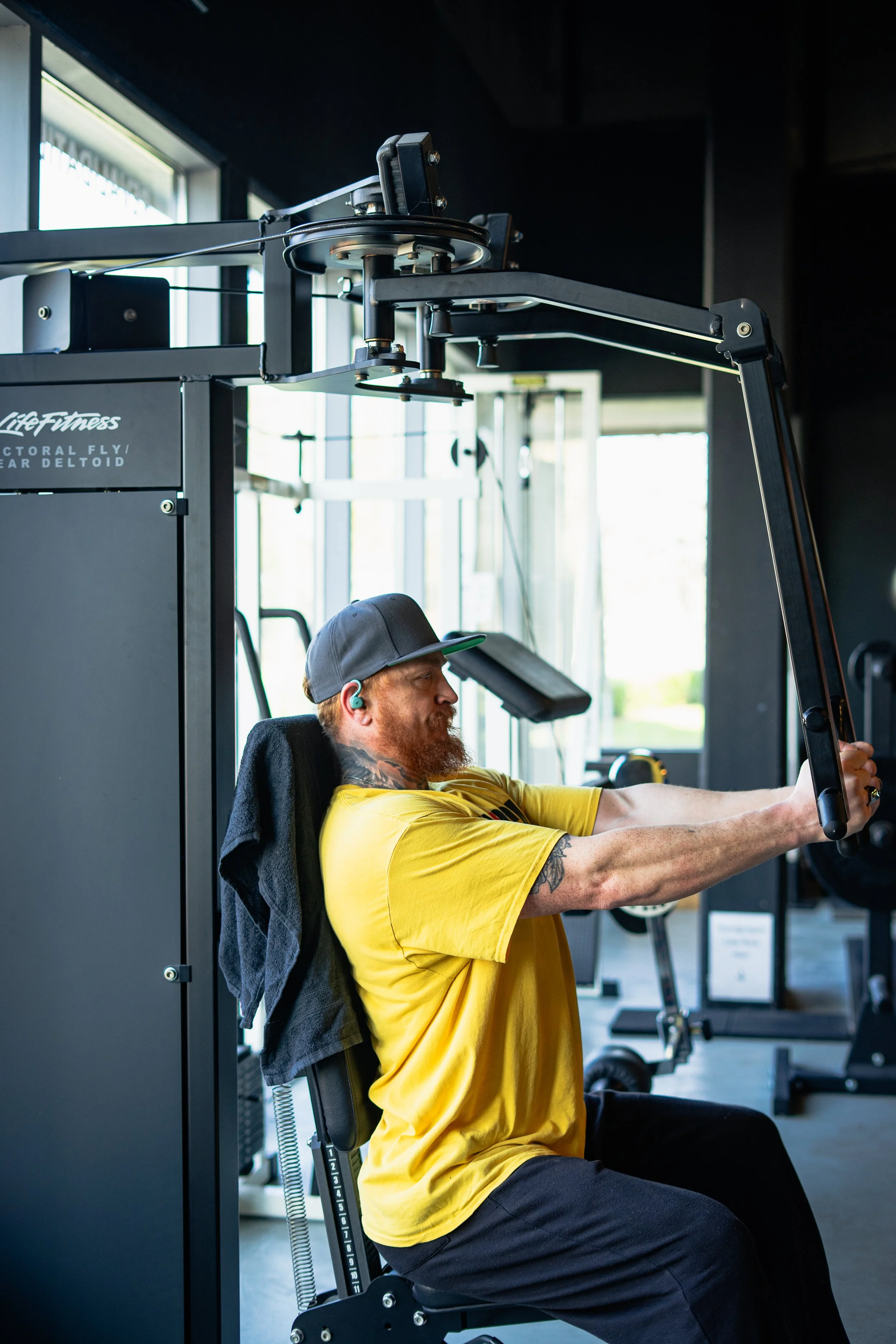 Man in een sportschool gebruikt een fitnessapparaat, met een zwart shirt en een baseballcap. Sportschool Emmen. Krachttraining.