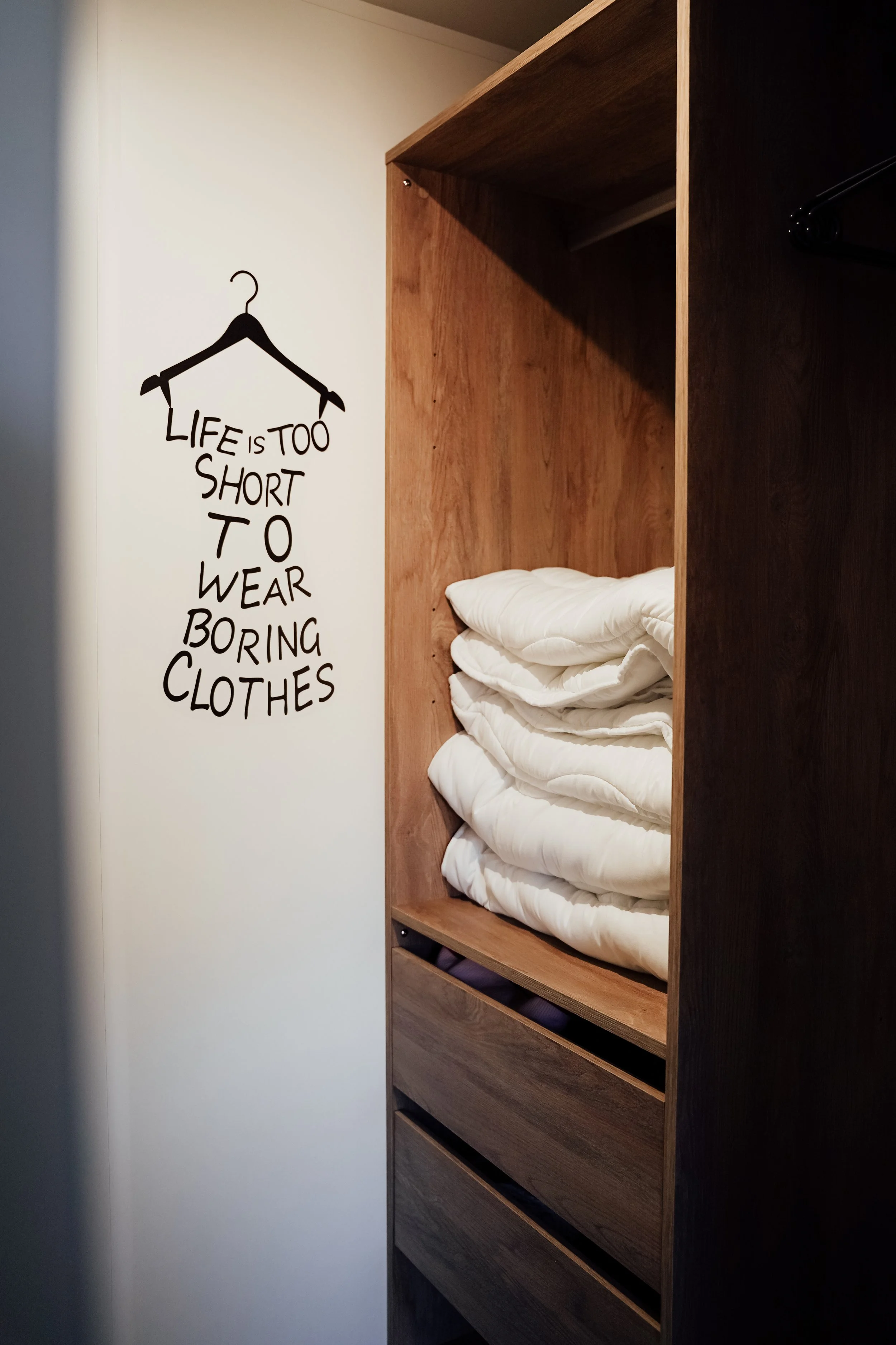 Een houten kast met gevouwen witte dekbedden en een muur met quote: "Life is too short to wear boring clothes". Vakantiepark. Chalet.