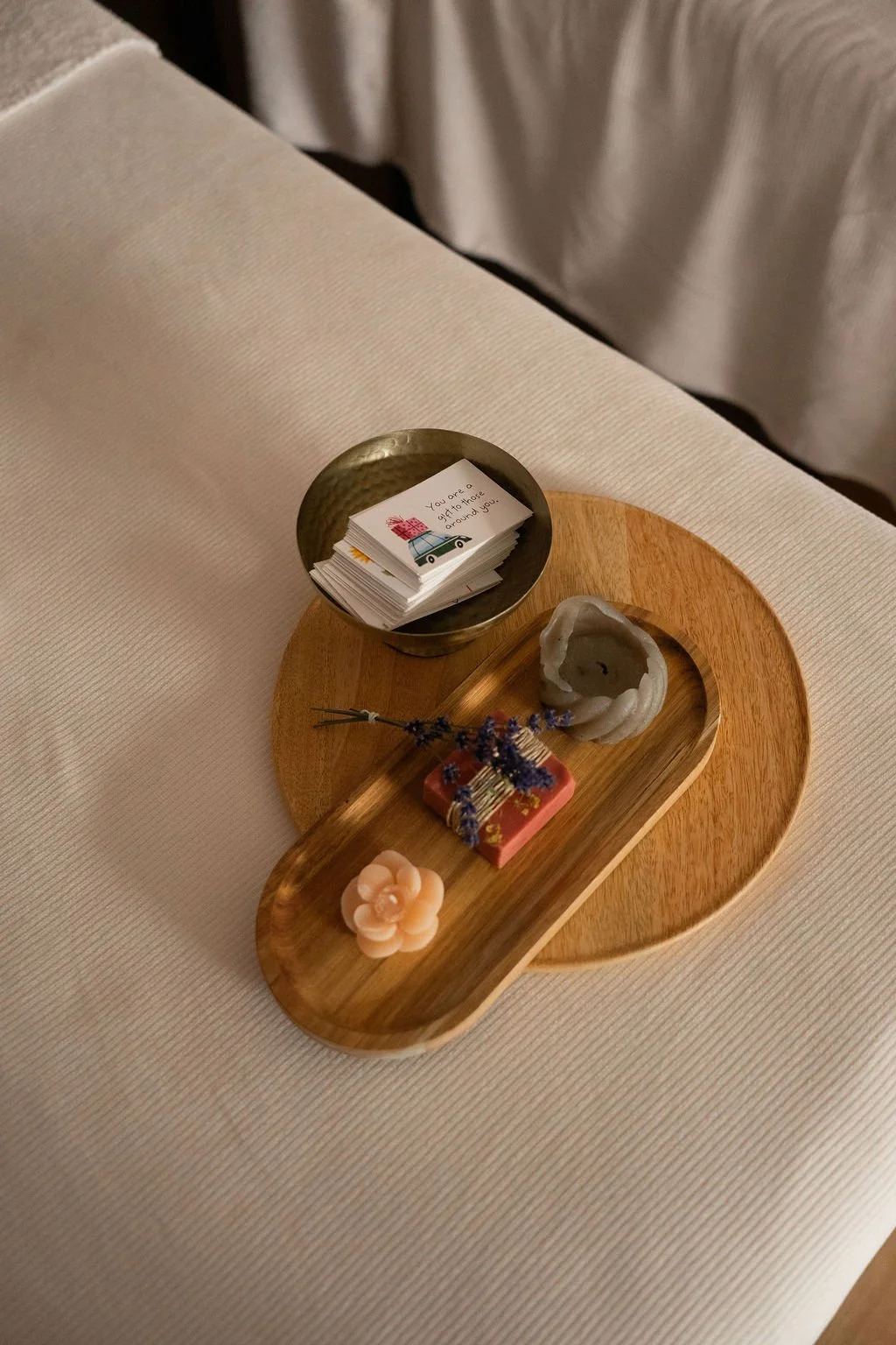 Een houten dienblad met decoratieve objecten, inclusief een kaars, lavendel bloemen, een klein cadeau en een stapel ansichtkaarten, op een effen tafel.