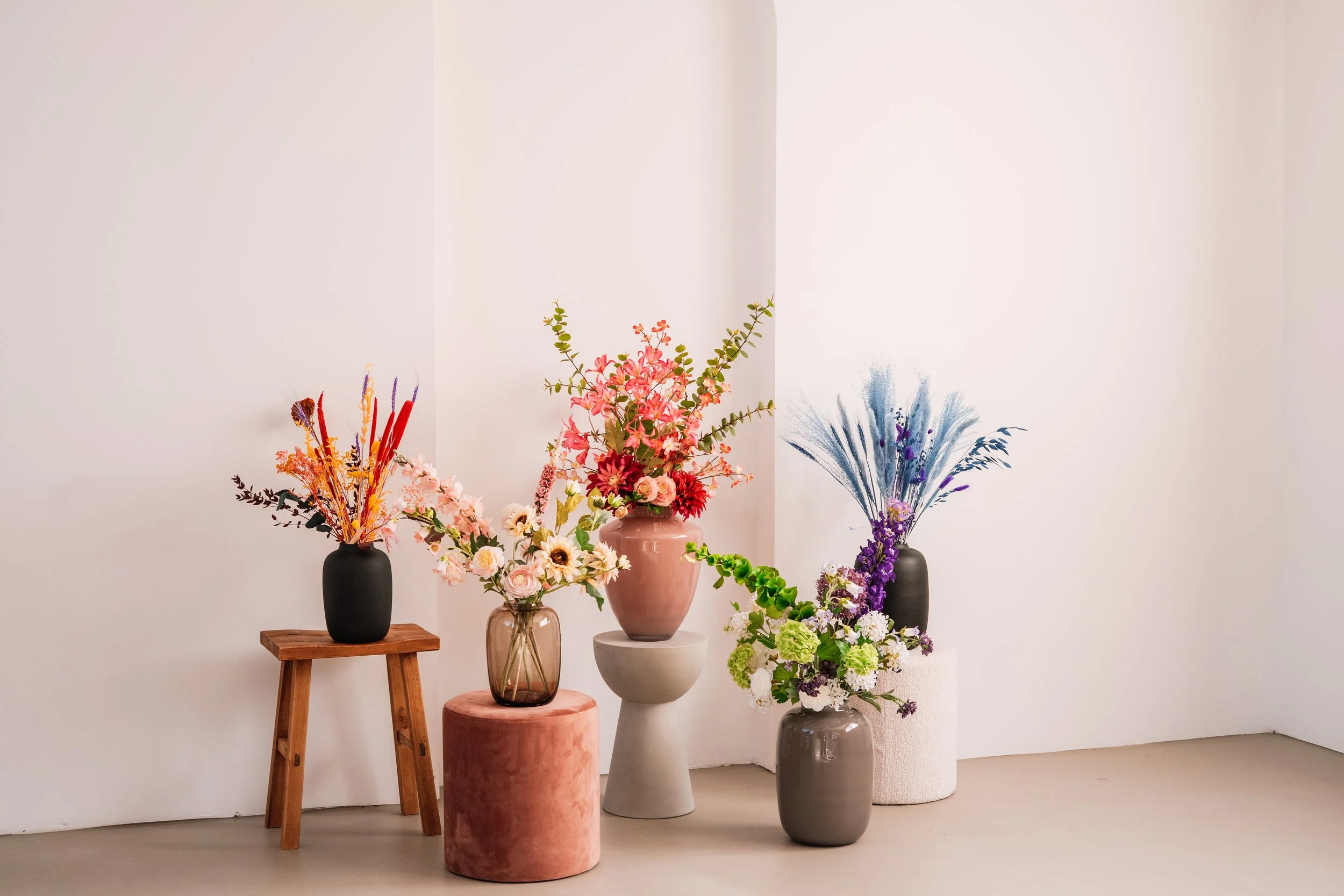 Vijf vazen met kleurrijke bloemen, geplaatst op verschillende onderstellen en een witte muur op de achtergrond. Floweris