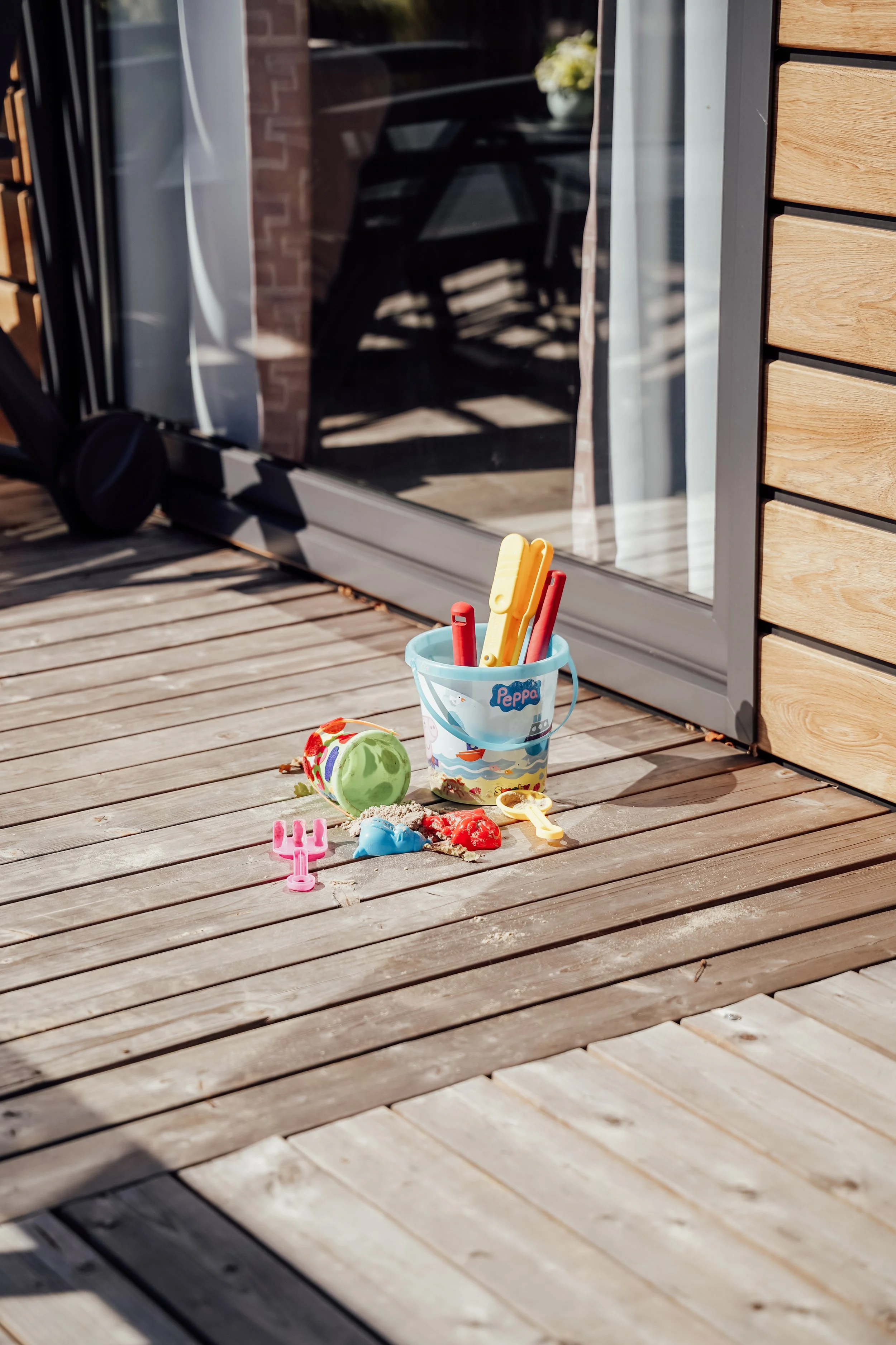 Speelgoed en zand op een houten terras buiten, met een glazen deur en een spiegeling van een stoel in de achtergrond.