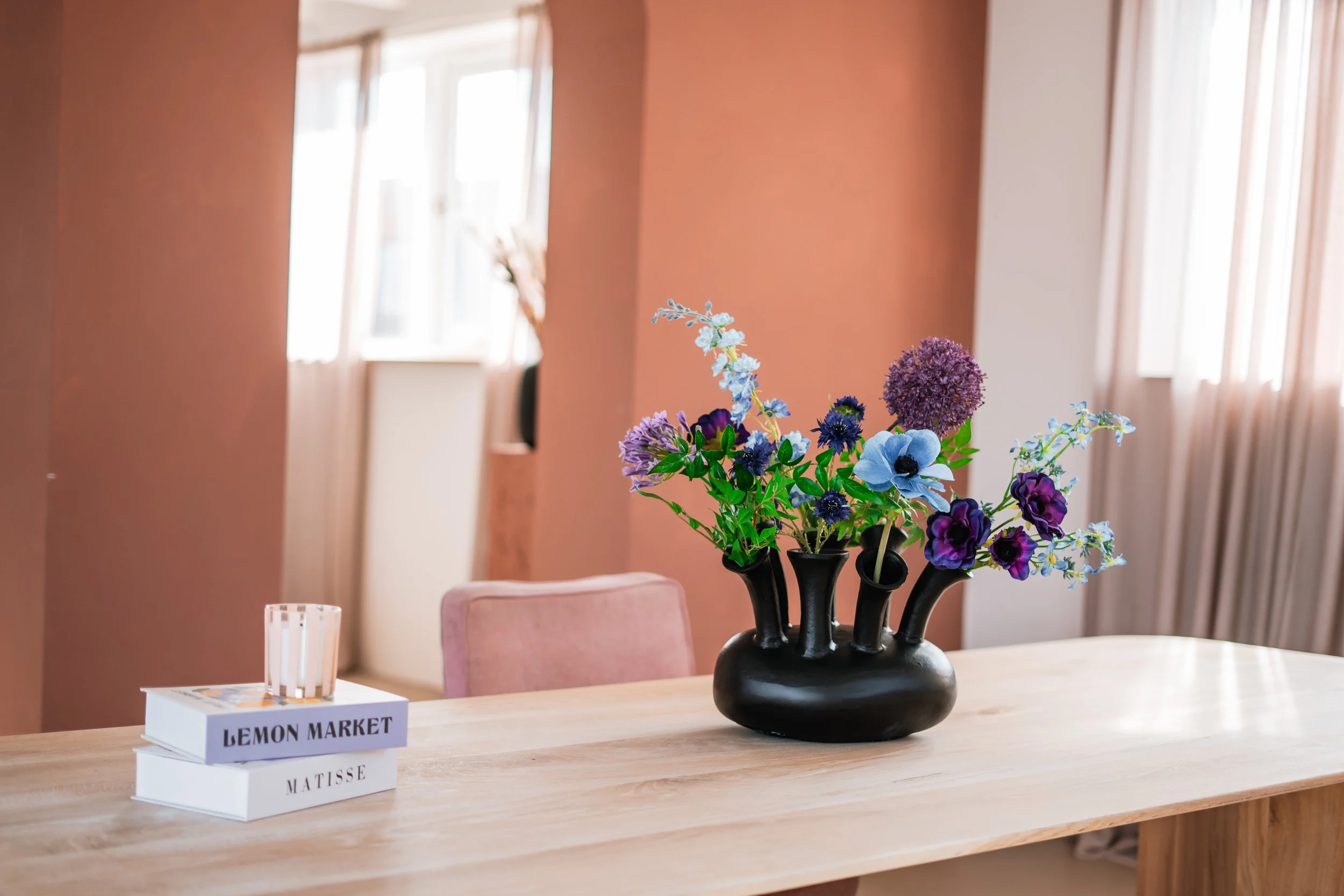 Decoratieve vaas met bloemen op een houten tafel, in een lichte kamer met ramen en roze muren. Floweris