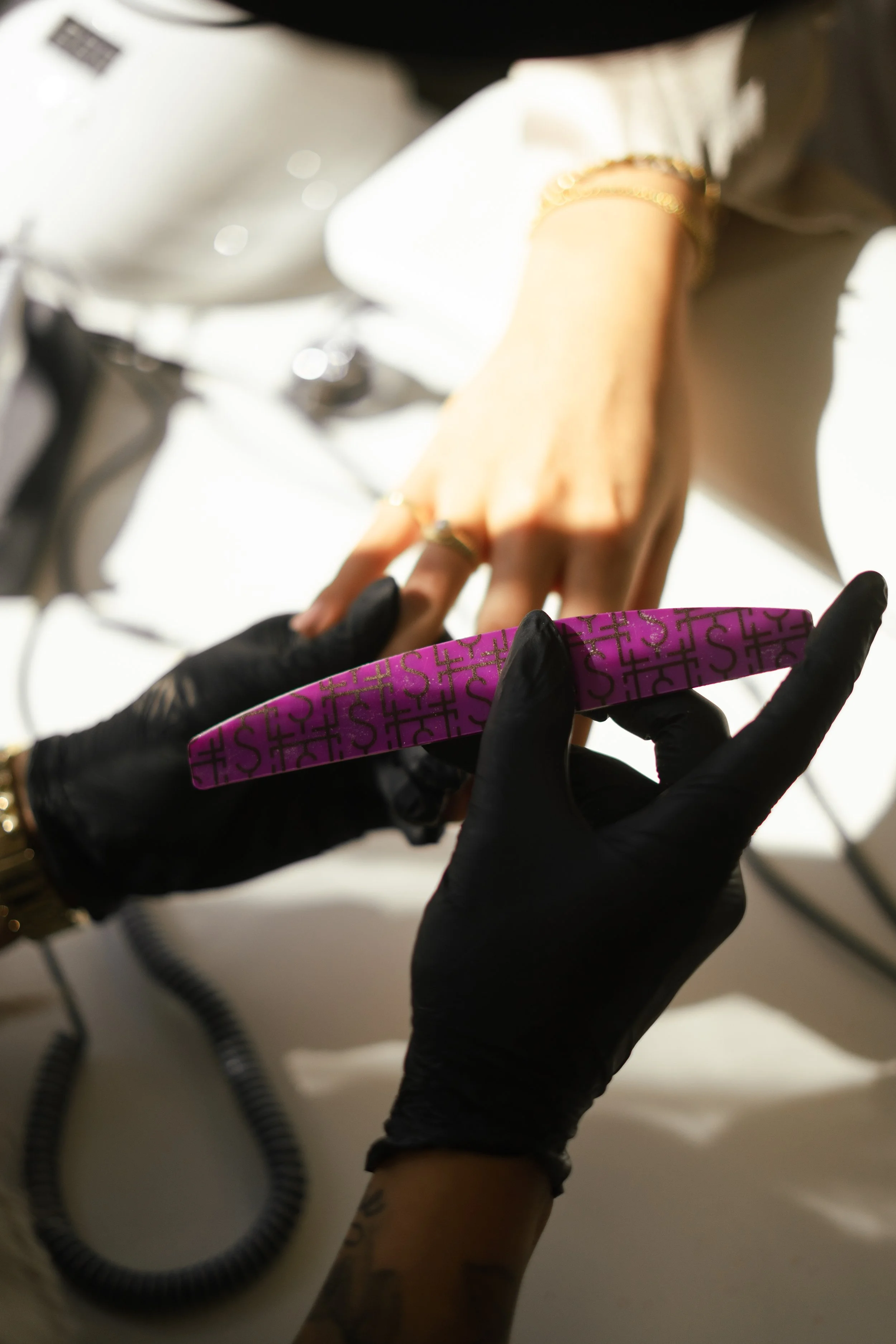 Persoon krijgt een nagellakbehandeling, met een hand die een zwarte handschoen draagt en een andere hand met sieraden. Een persoon in de achtergrond met een lichte blouse en een gouden armband.