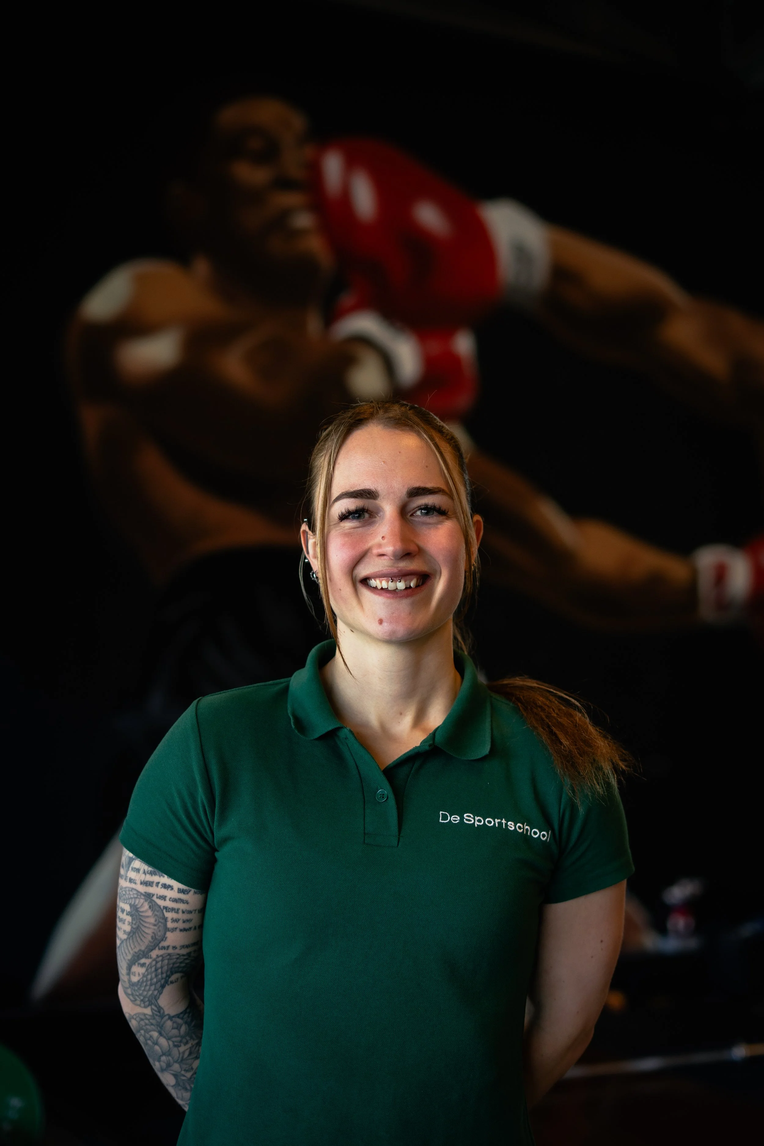Een glimlachende vrouw in een groene polo met de tekst 'De Sportschool', staand voor een achtergrond met een grote afbeelding van een bokser die uitrust na een wedstrijd.