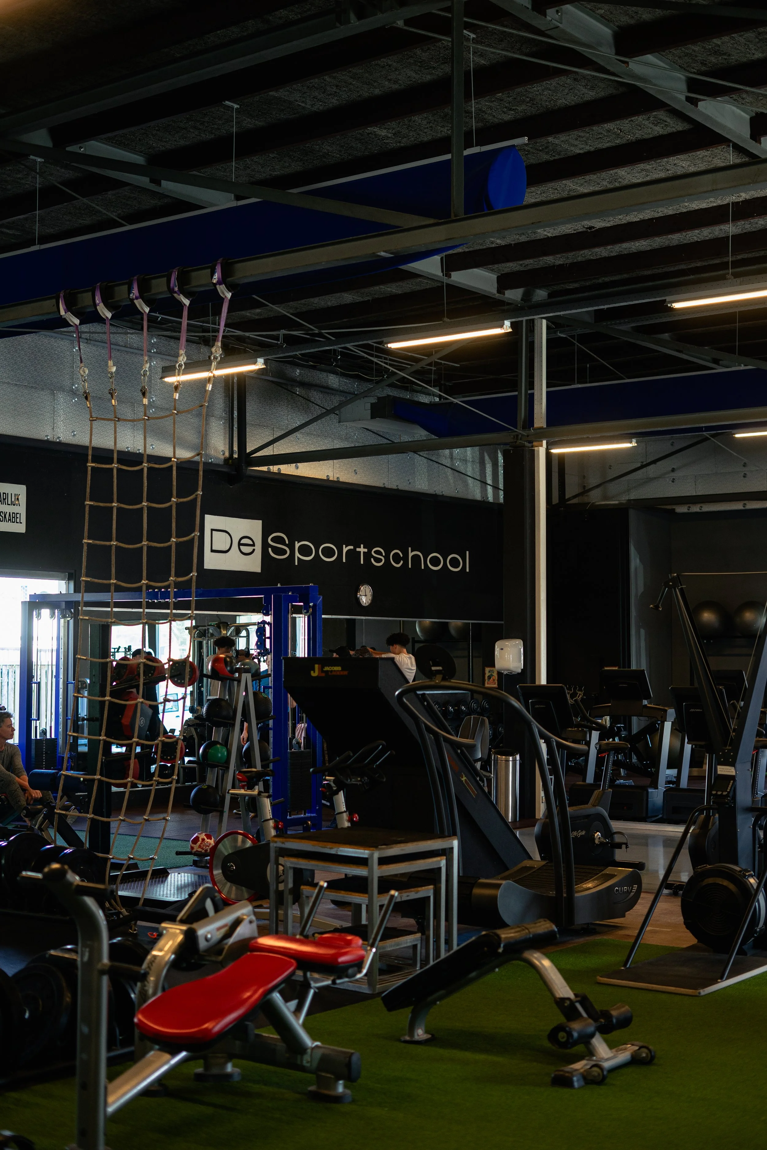 Binnenruimte van een sportschool met fitnessapparatuur, een klimrek, en sporters die trainen. Sportschool Emmen. Krachttraining.