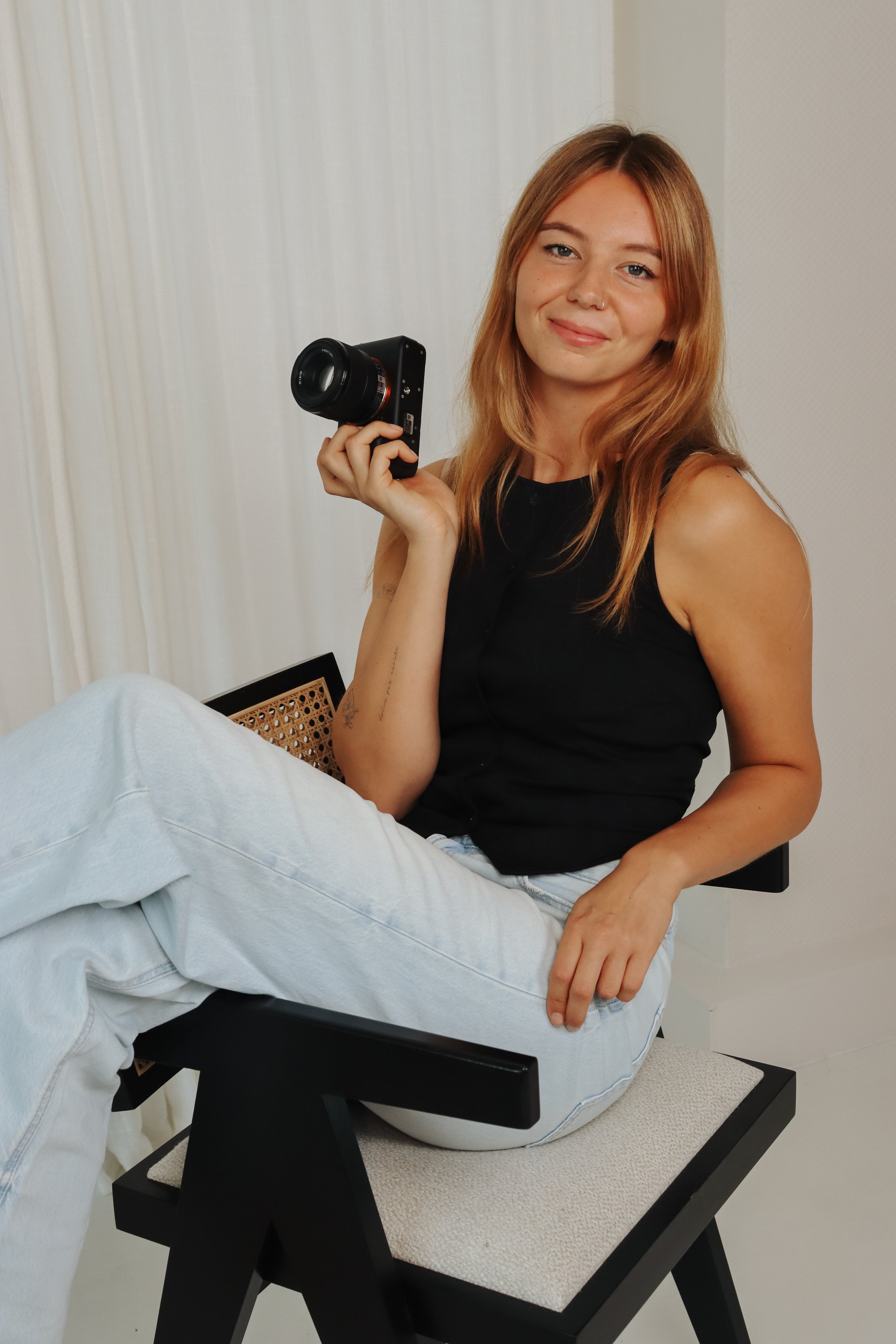 Vrouw met rode haar, zittend op een stoel, die een kleine camera vasthoudt en glimlacht.