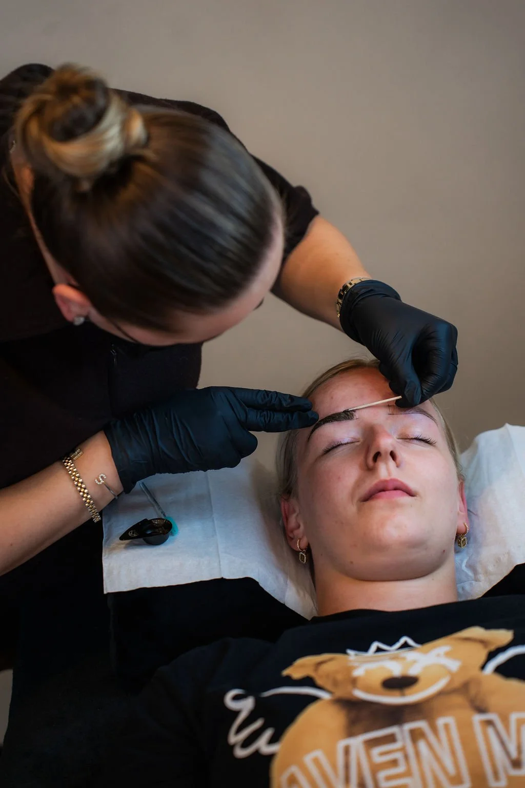 Vrouw ligt met gesloten ogen op een behandeltafel terwijl een specialist haar wenkbrauwen tattooers met een pincet en een pincet. Beautysalon.