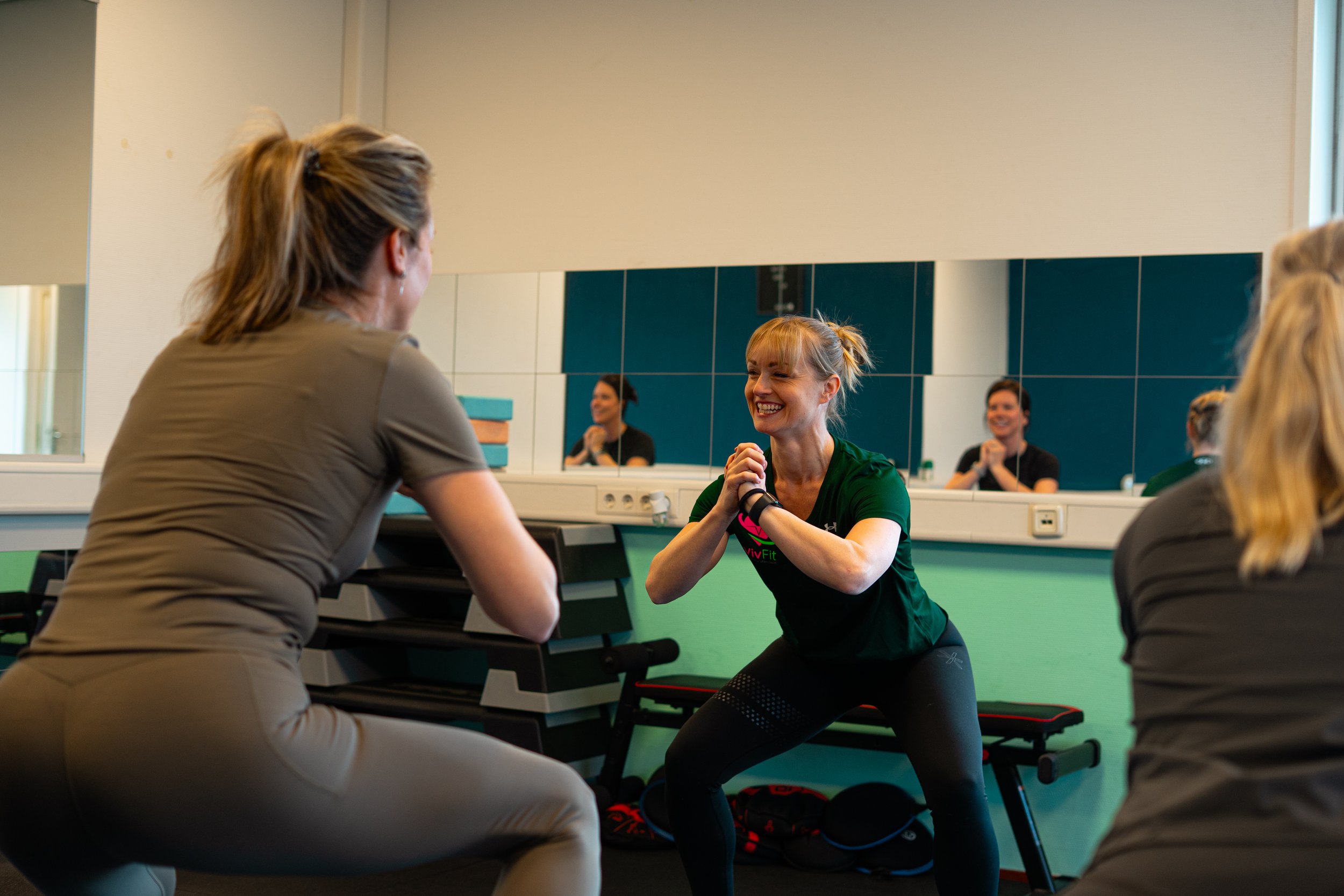 Vrouw die squats doet tijdens een fitnessles, lachend en in een groene T-shirt, met instructrices die haar begeleiden in een sportschool met spiegels op de muur. Sportschool. Fit worden. Content creatie MCC Agency.