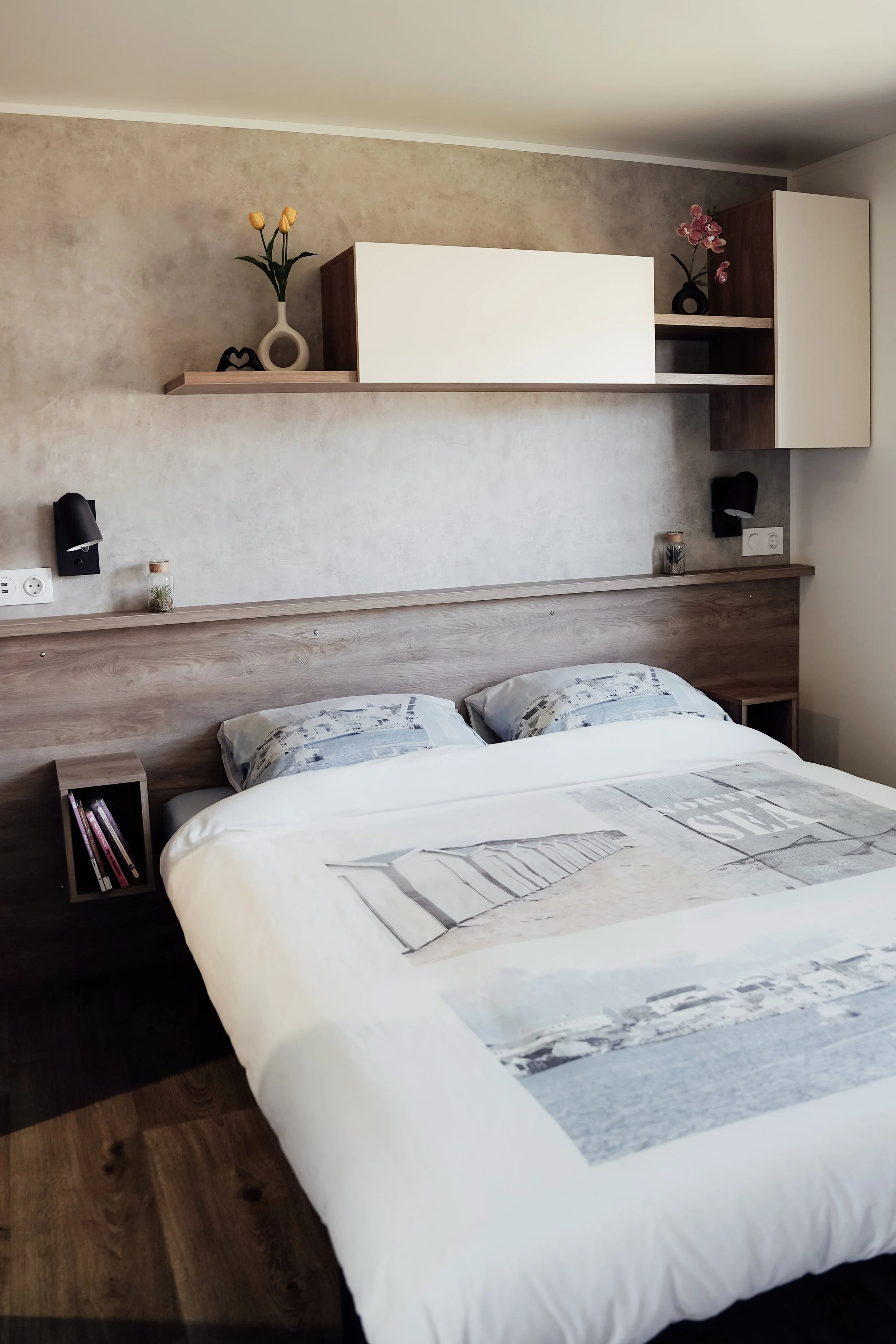 Een slaapkamer met een bed, een houten hoofdeinde, ingebouwde leeslampen, planken boven het bed met vazen en decoraties, en een muur met een subtiele textuur. Vakantiepark. Chalet.