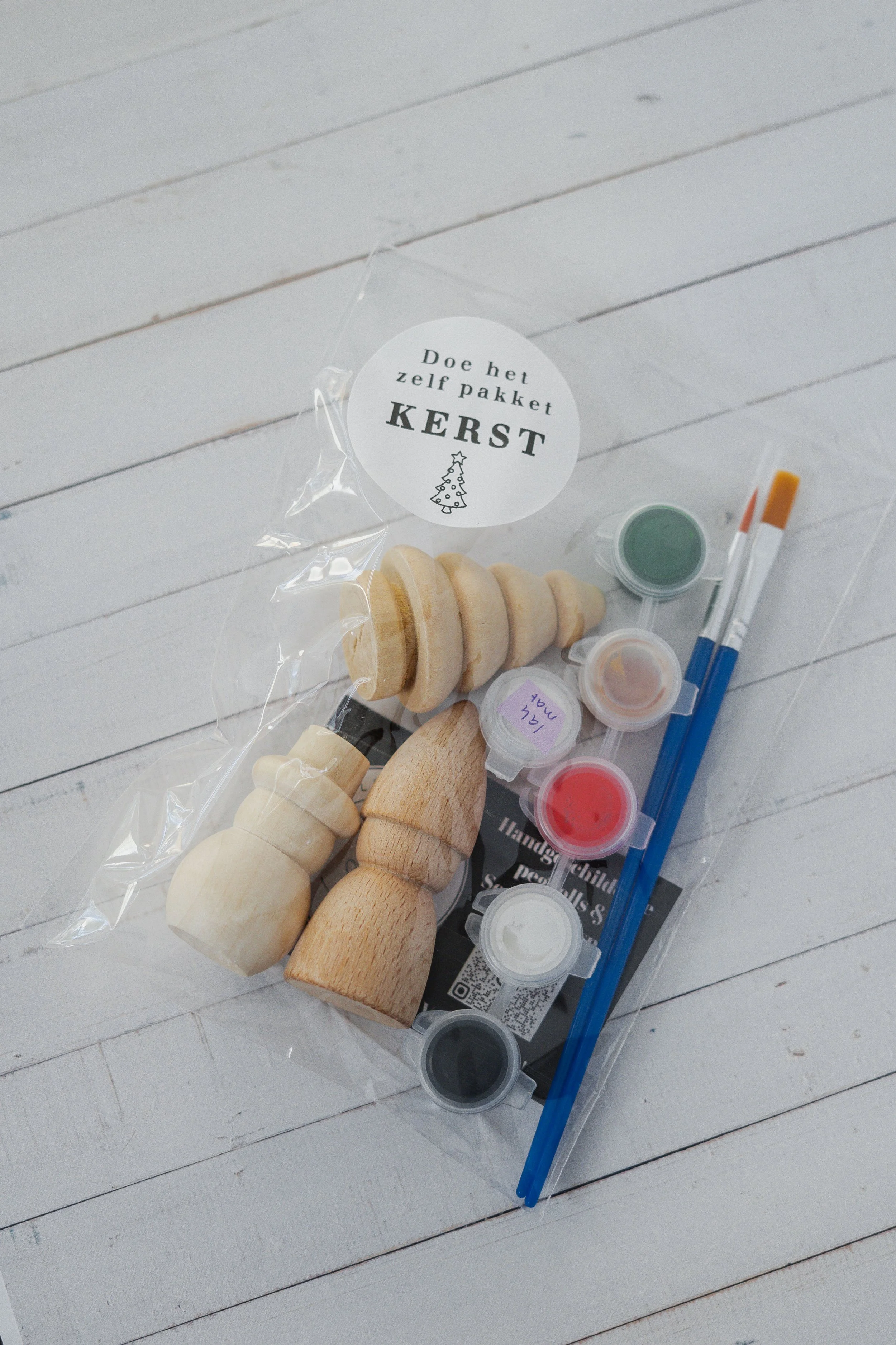 Knutselpakket met houten kerstfiguren, verf en penselen op een witte houten tafel.