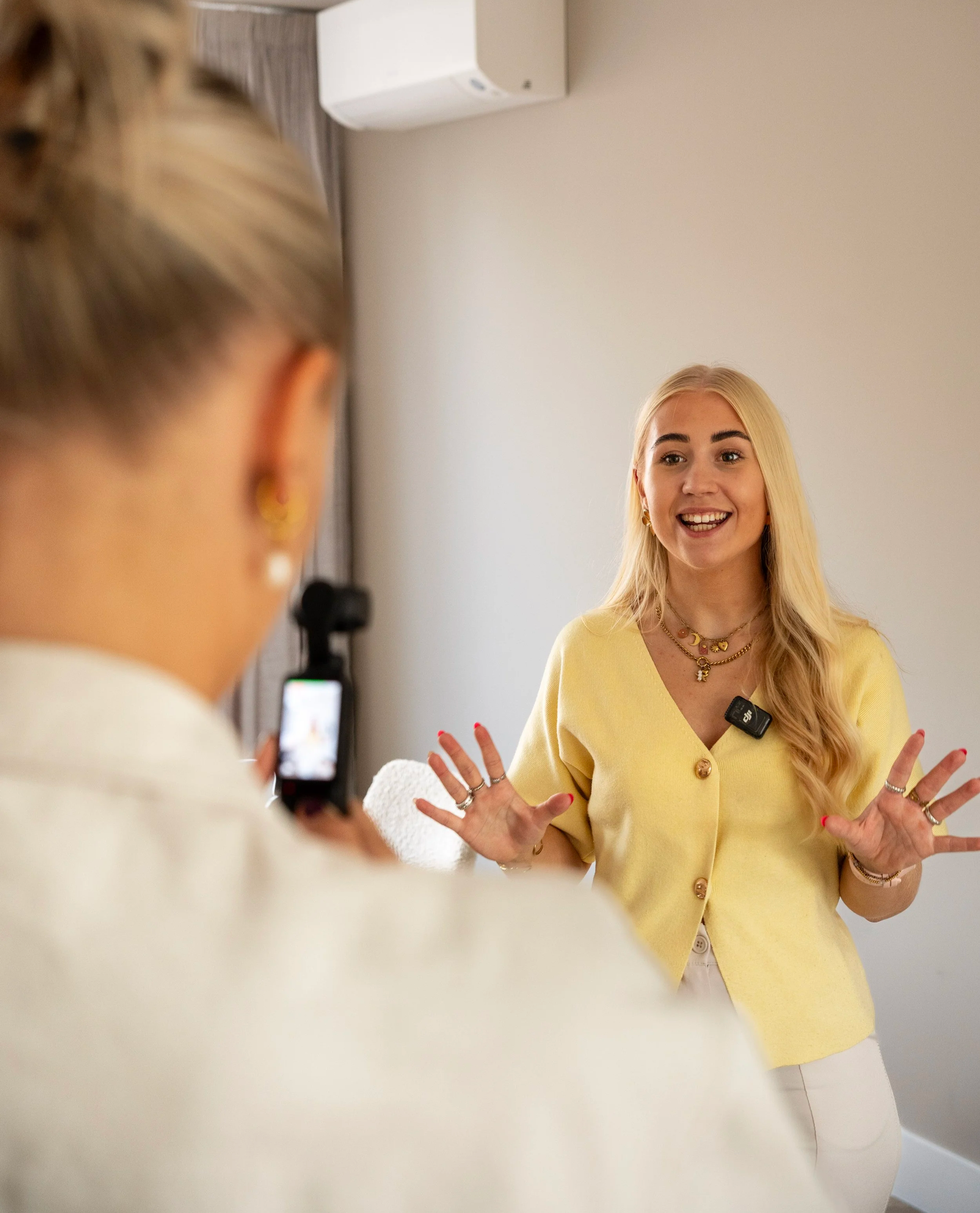 Vrouw in gele top geeft interview en wordt gefilmd door persoon met telefoon.