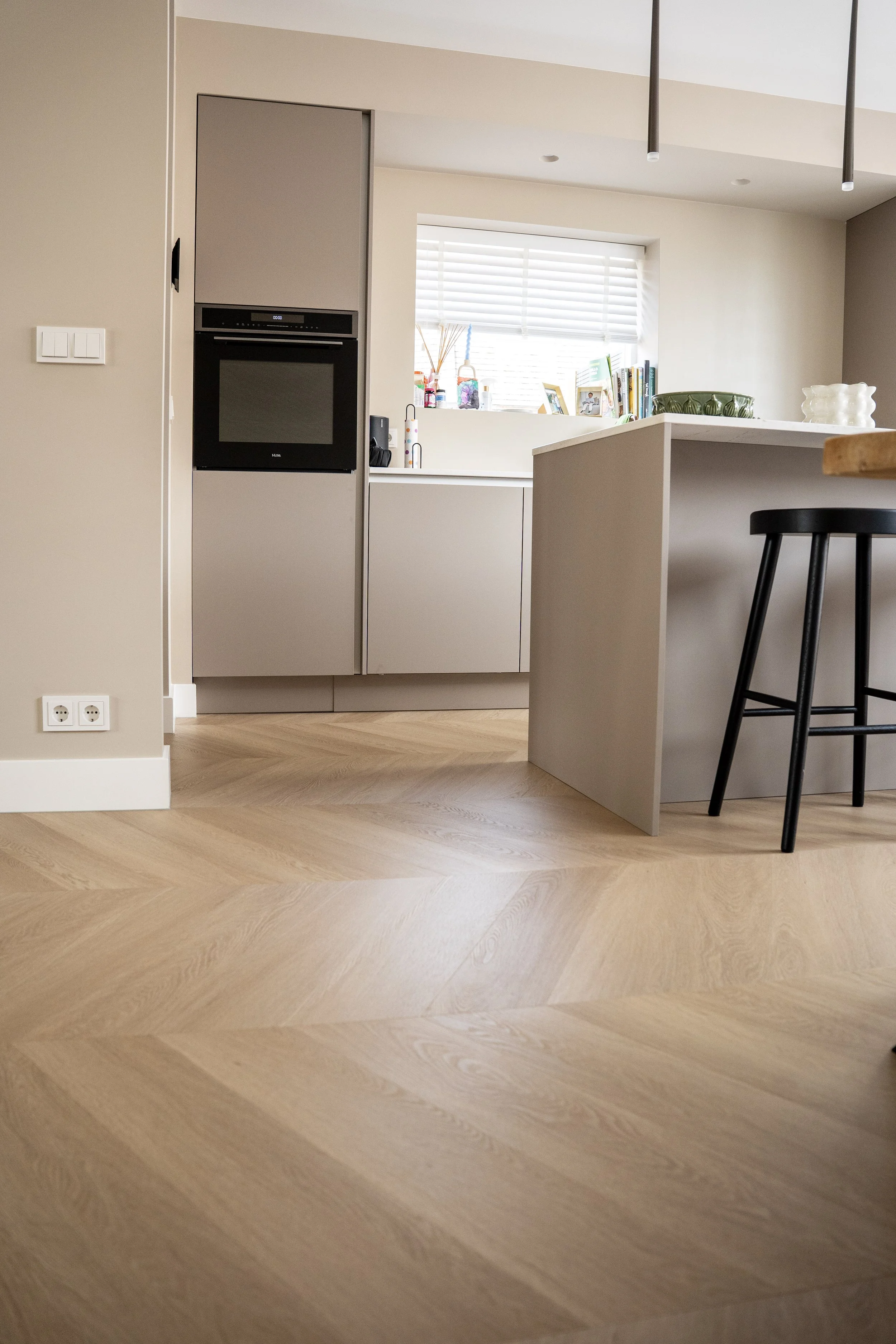 Een modern keuken met een houten vloer, een stuur met vier poten en een keukenblok met verschillende items en decoraties. Een raam met witte jaloezieën op de achtergrond. Laminaatpaleis Ede