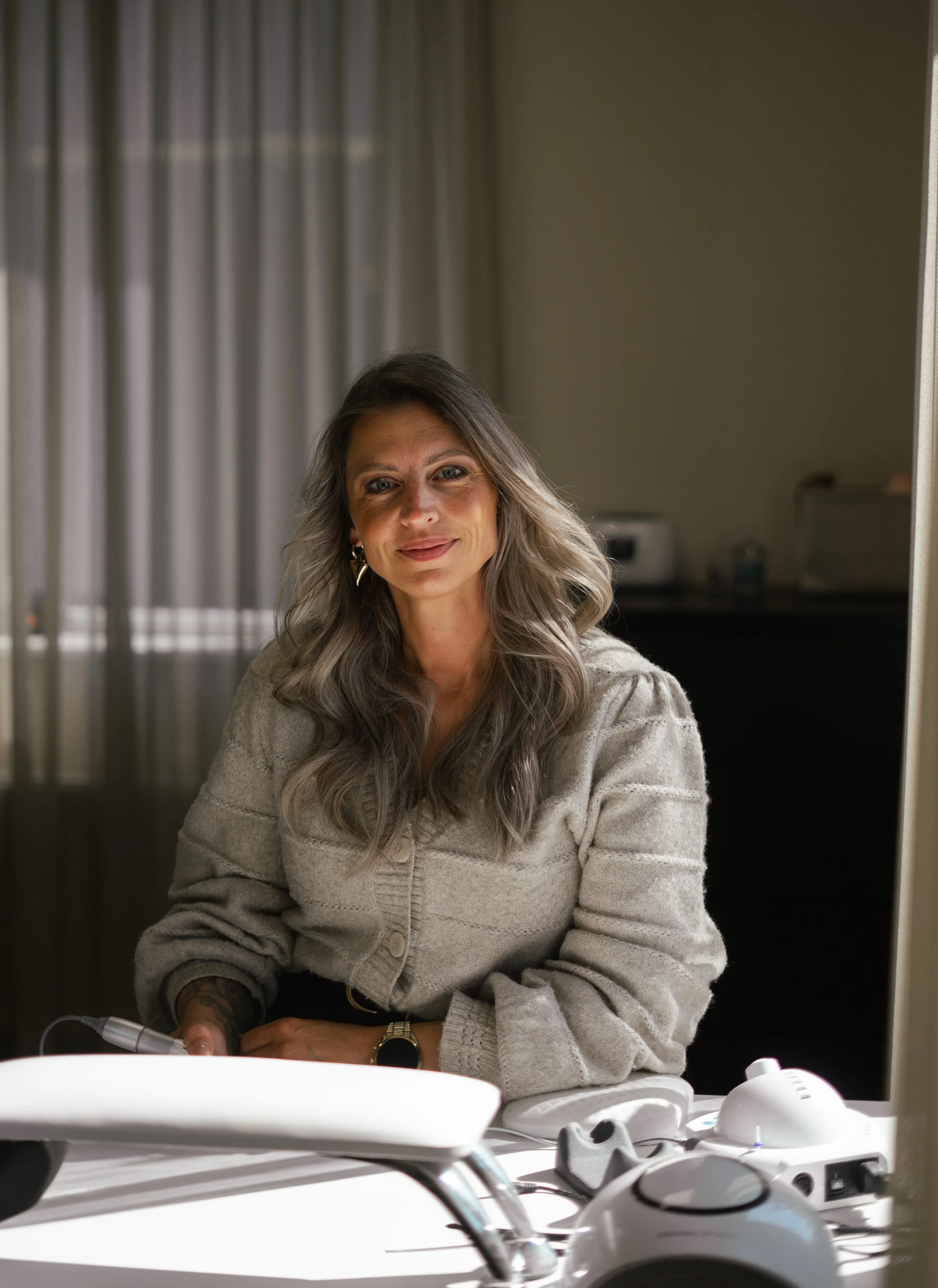 Vrouw met grijze haar en een beige trui zittend bij een tafel met medische apparatuur, in een kamer met gordijnen op de achtergrond.