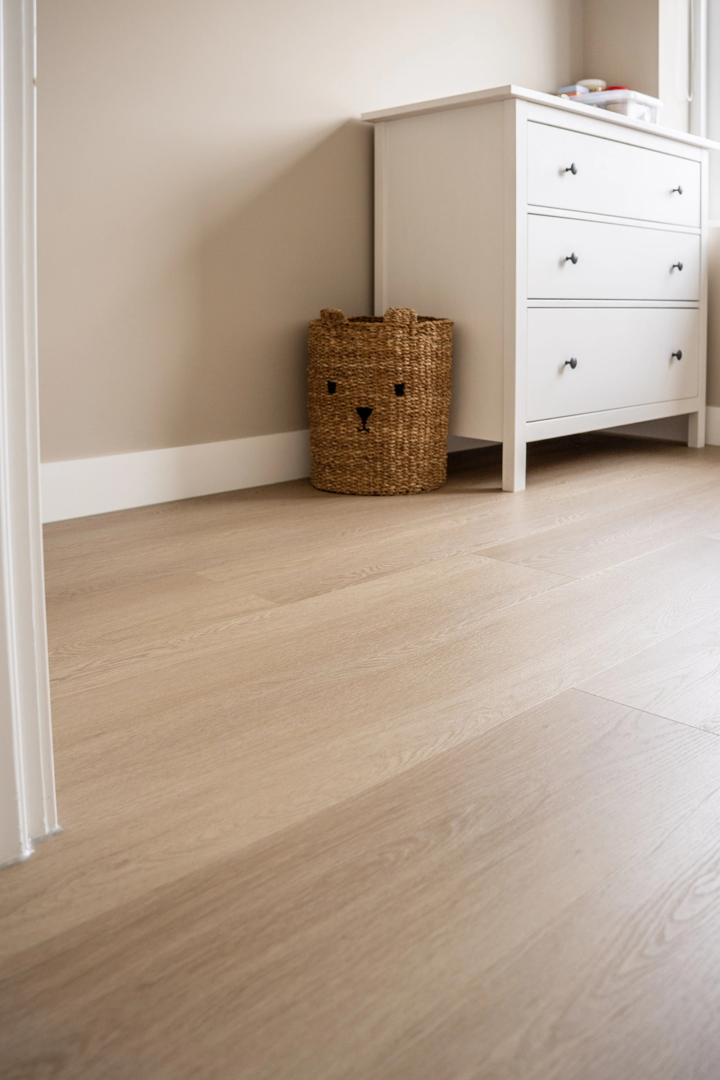 Een kamerhoek met een houten vloer, een witte ladekast en een bamboe mand met een hondenkop opzet. Laminaatpaleis Ede