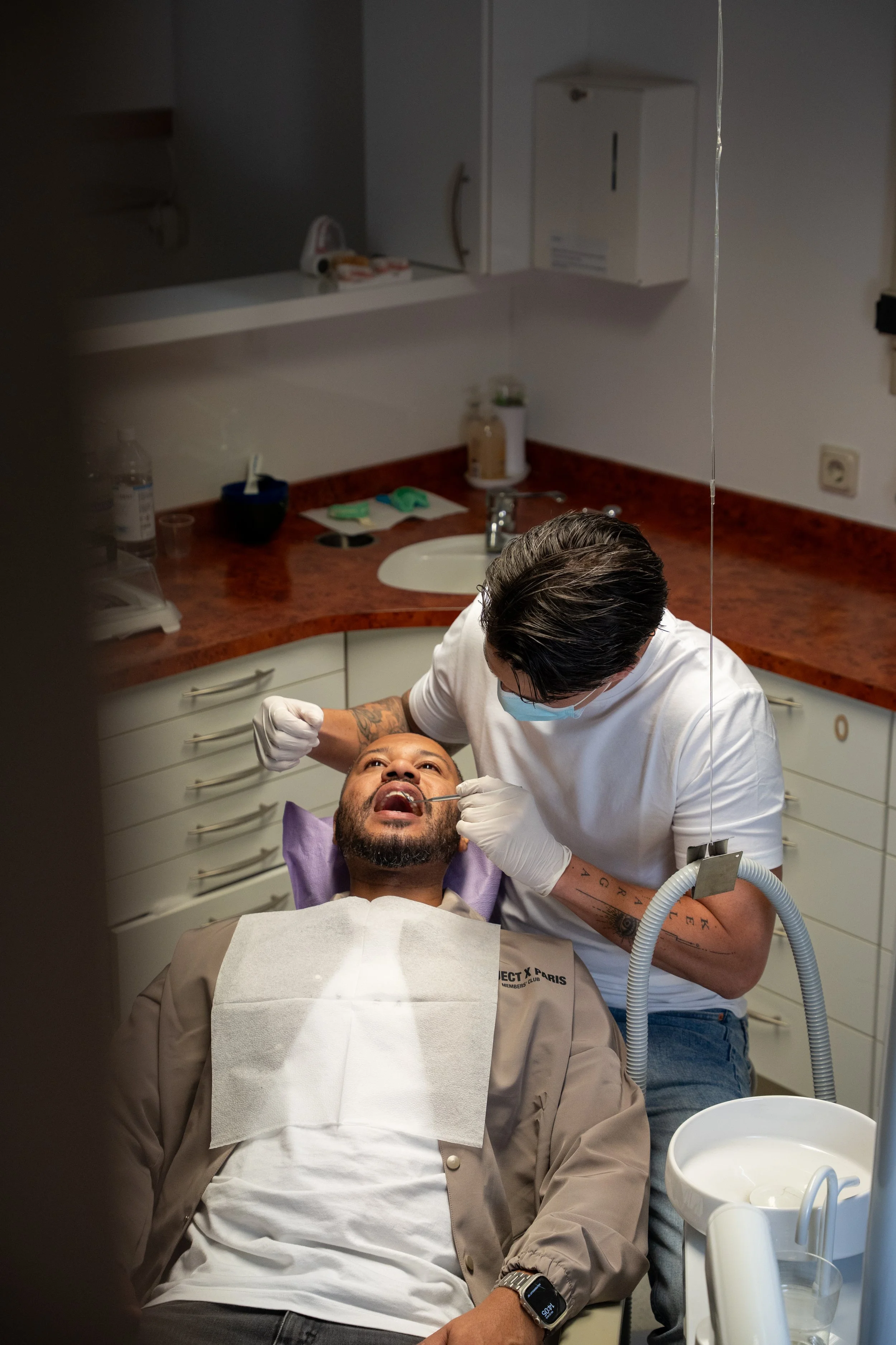 Een man ligt achterover in een tandartsstoel terwijl een tandarts of arts een behandeling uitvoert in een kliniek met witte kasten en een rood/oranje werkblad. Wittewel. Tandenbleken.