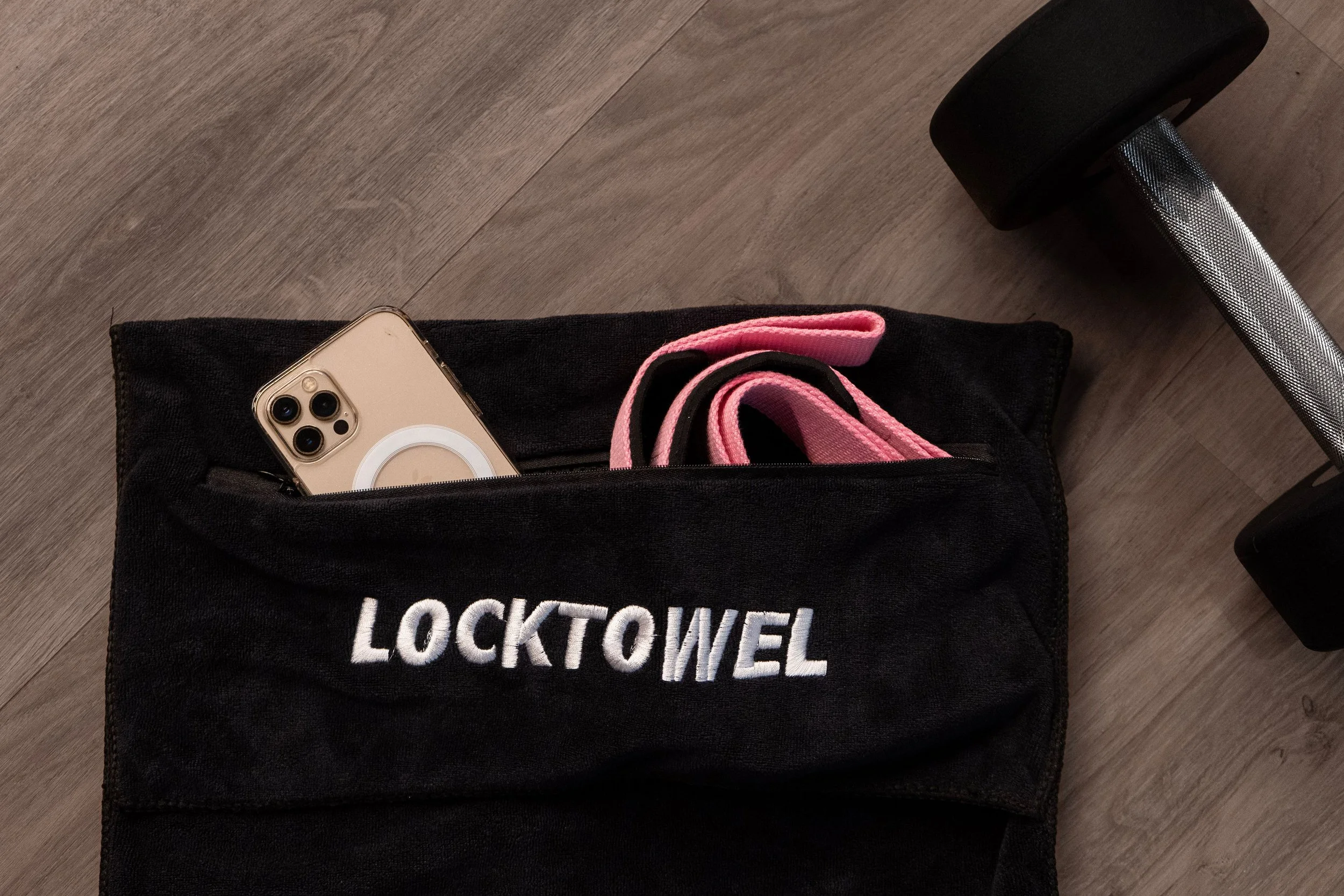 Zwarte sporttas met de tekst 'LOCKTOWEL', een smartphone met oplader, pinke sporthanddoek en een halter aan de vloer. Sporthanddoek. Locktowel.