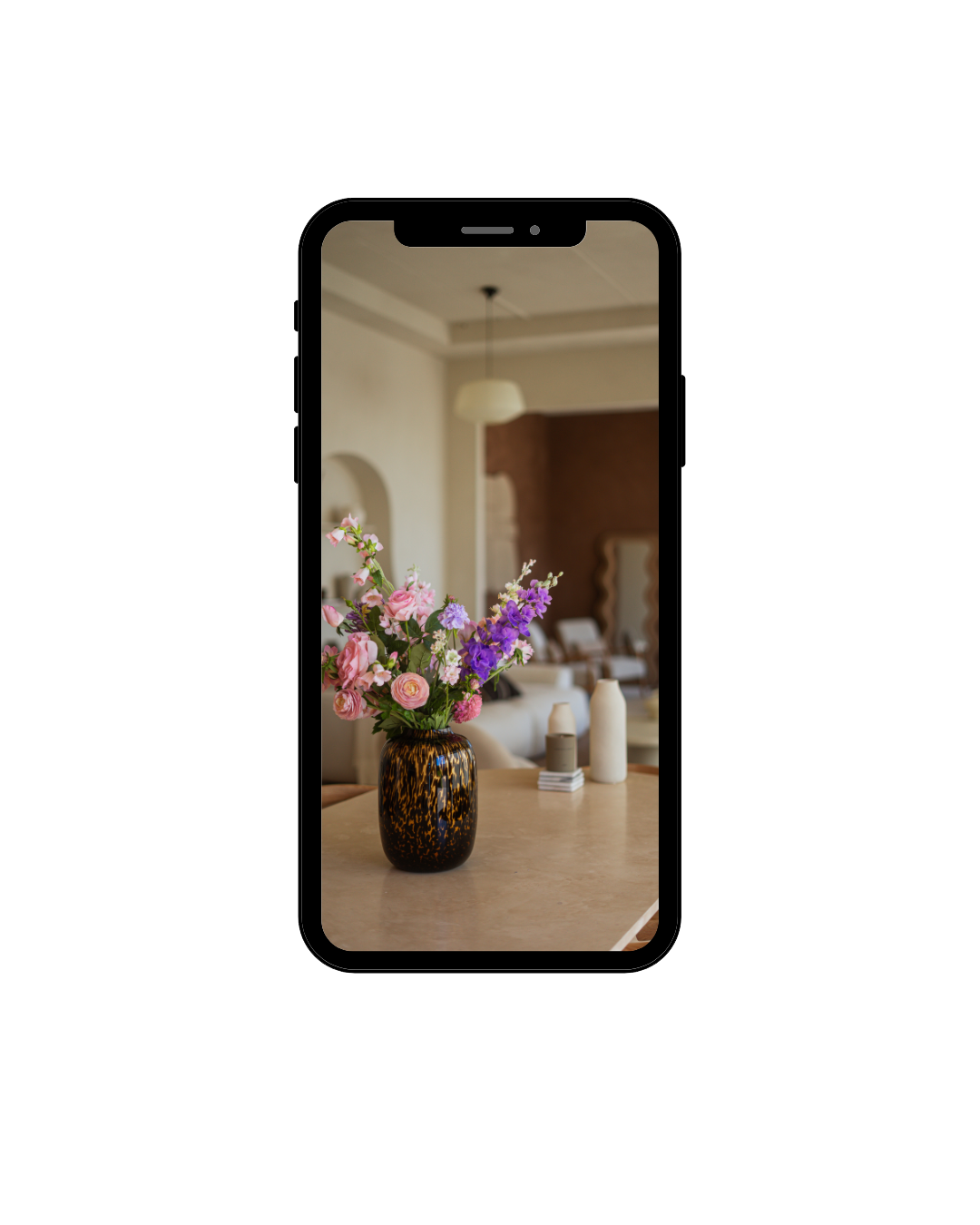 Een smartphone met een foto van een bloemenvaas met roze en paarse bloemen op een houten tafel in een woonkamer.
