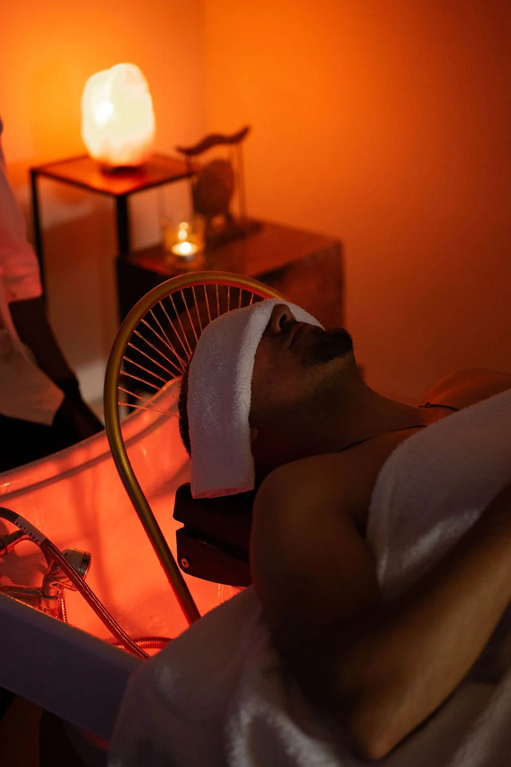 Man ligt ontspannen op een behandelbed met een witte handdoek op het hoofd in een spa of wellnessruimte met oranje verlichting. Japanse headspa massage. Selfcare moment