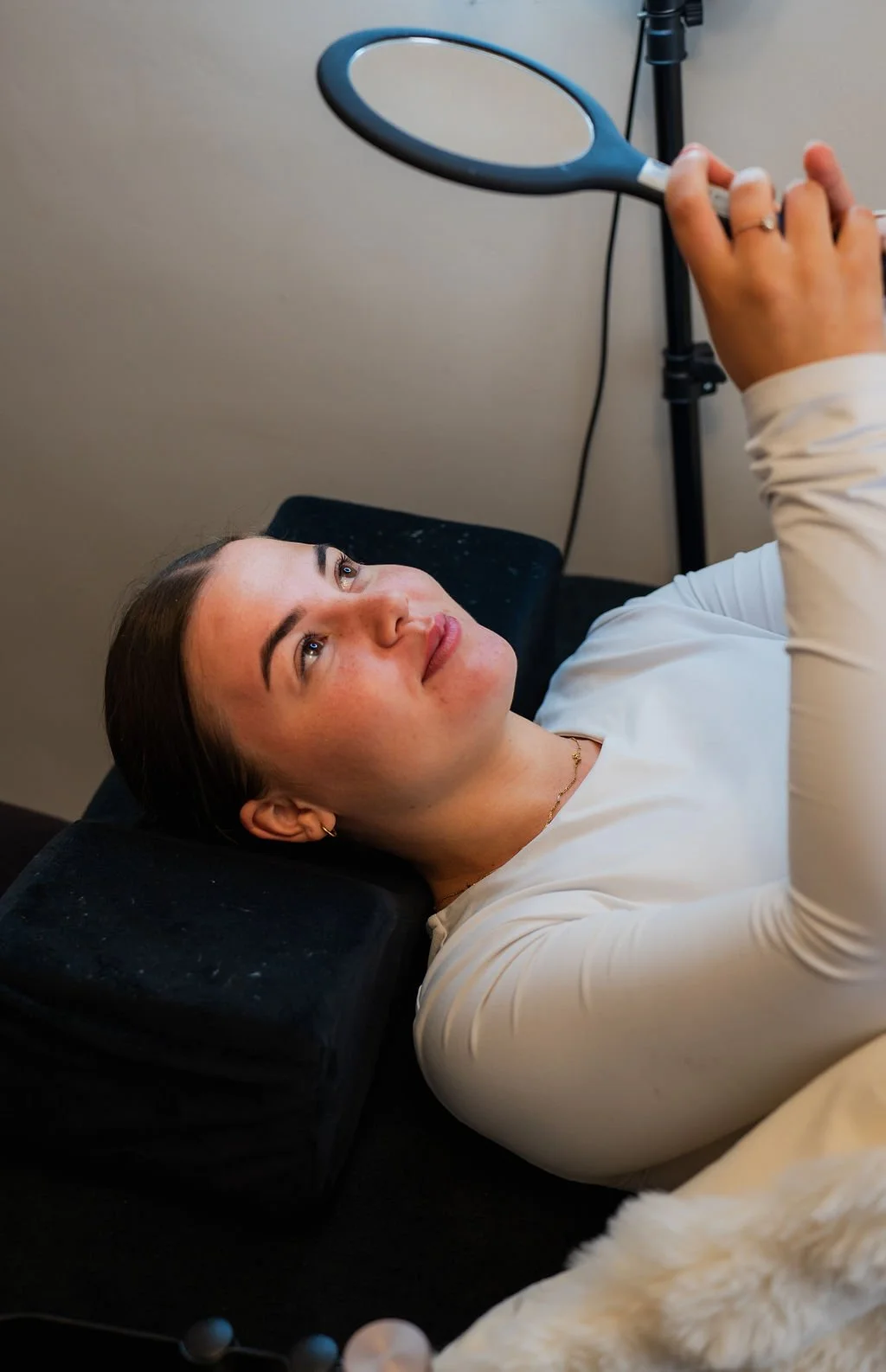 Vrouw ligt op een bankje met een hoofdsteun, kijkt omhoog naar een hand die een spiegel vasthoudt. Beautysalon.