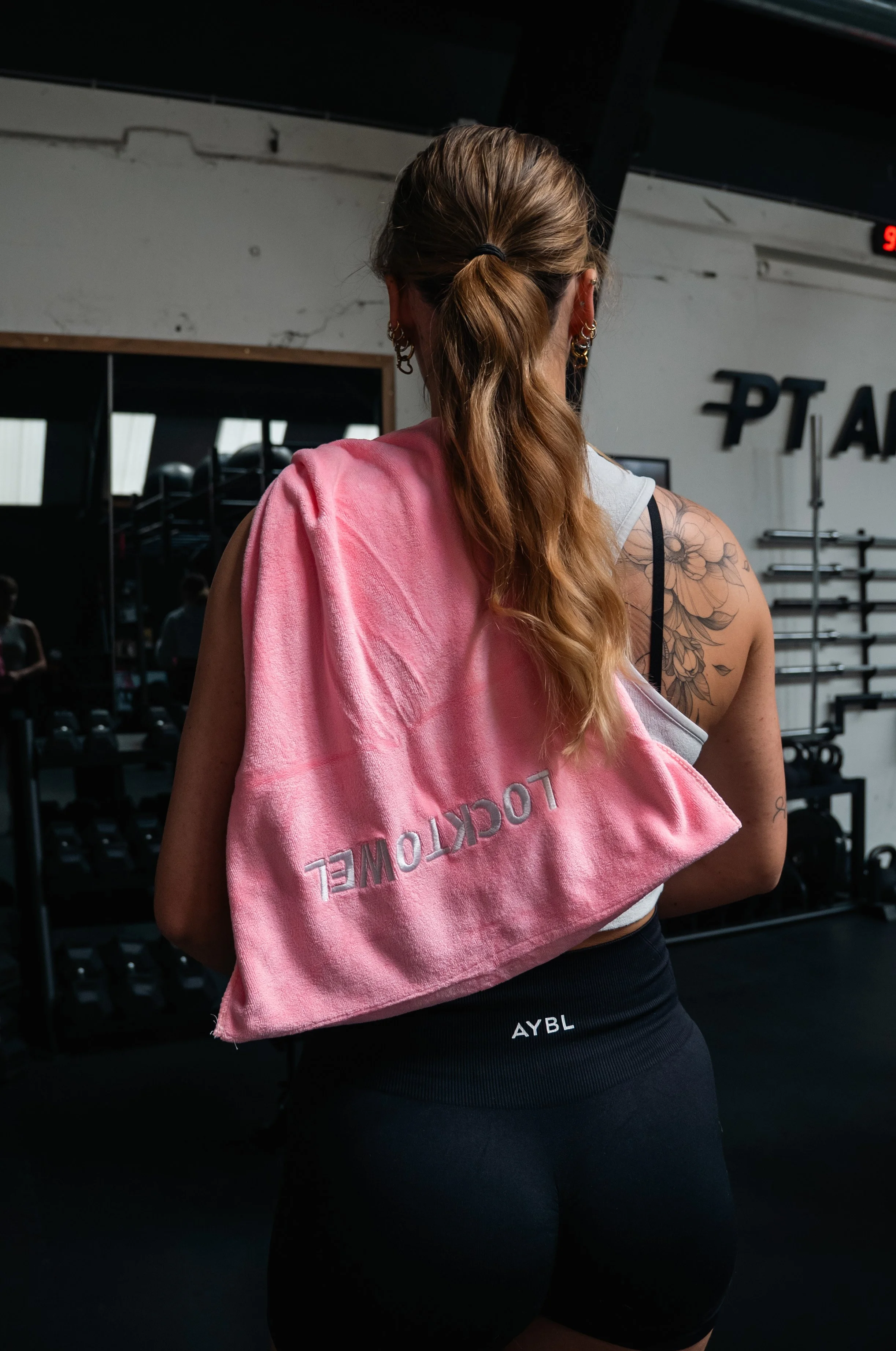 Vrouw met lang, golvend haar in een sportschool, draagt een wit sporttop, zwarte sportbroek met tekst 'AYBL', en heeft een roze handdoek met de tekst 'LOCKTOWEL' over haar schouder.
