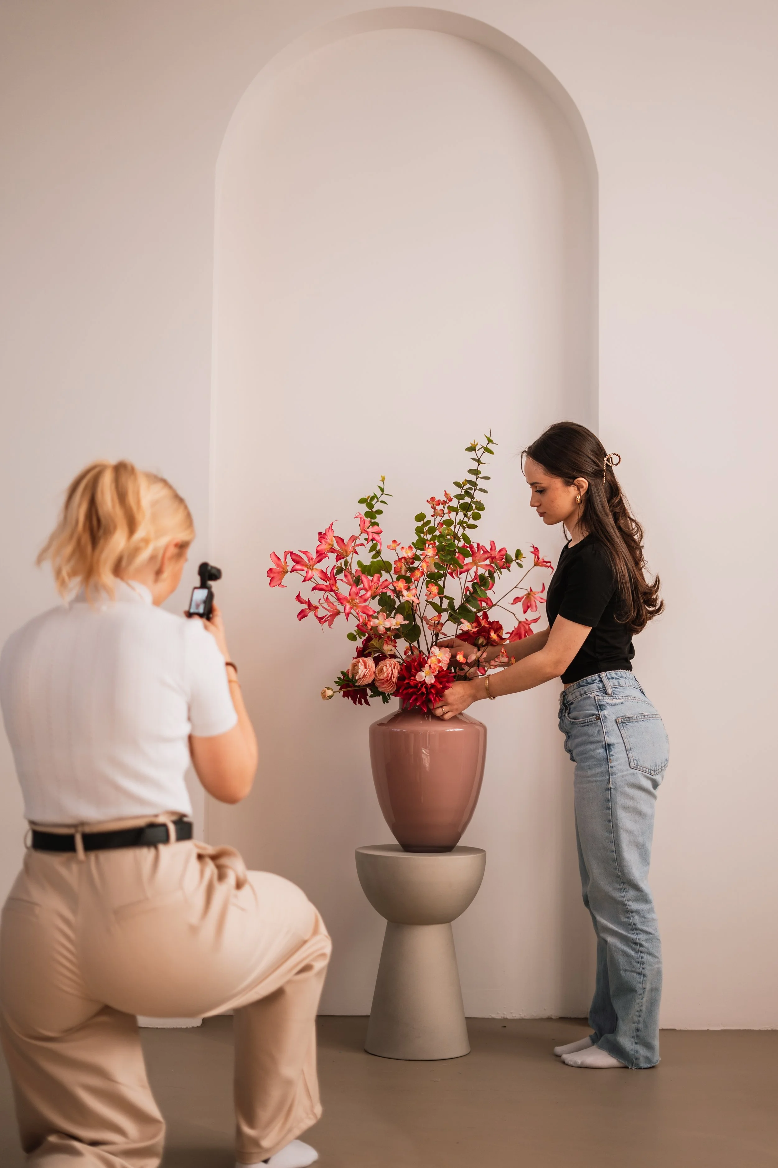 Twee vrouwen maken een foto van een bloemstuk in een grote vaas, die op een moderne losse tafel staat tegen een witte muur.