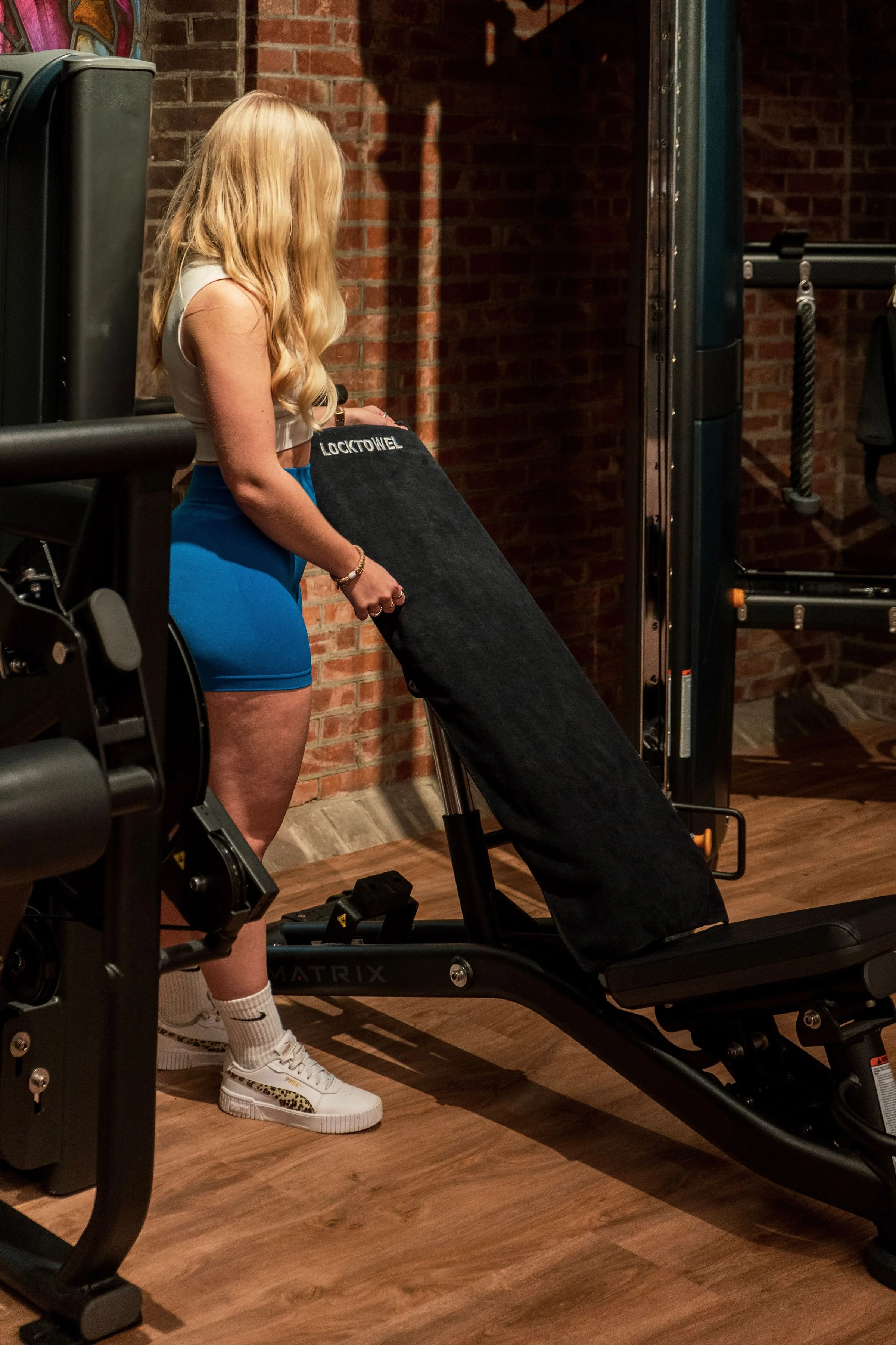 Een vrouw met lang blond haar, gekleed in een witte top en blauwe short, staat in een fitnessruimte en houdt eenStretch Apparatus. Sporthanddoek. Locktowel.