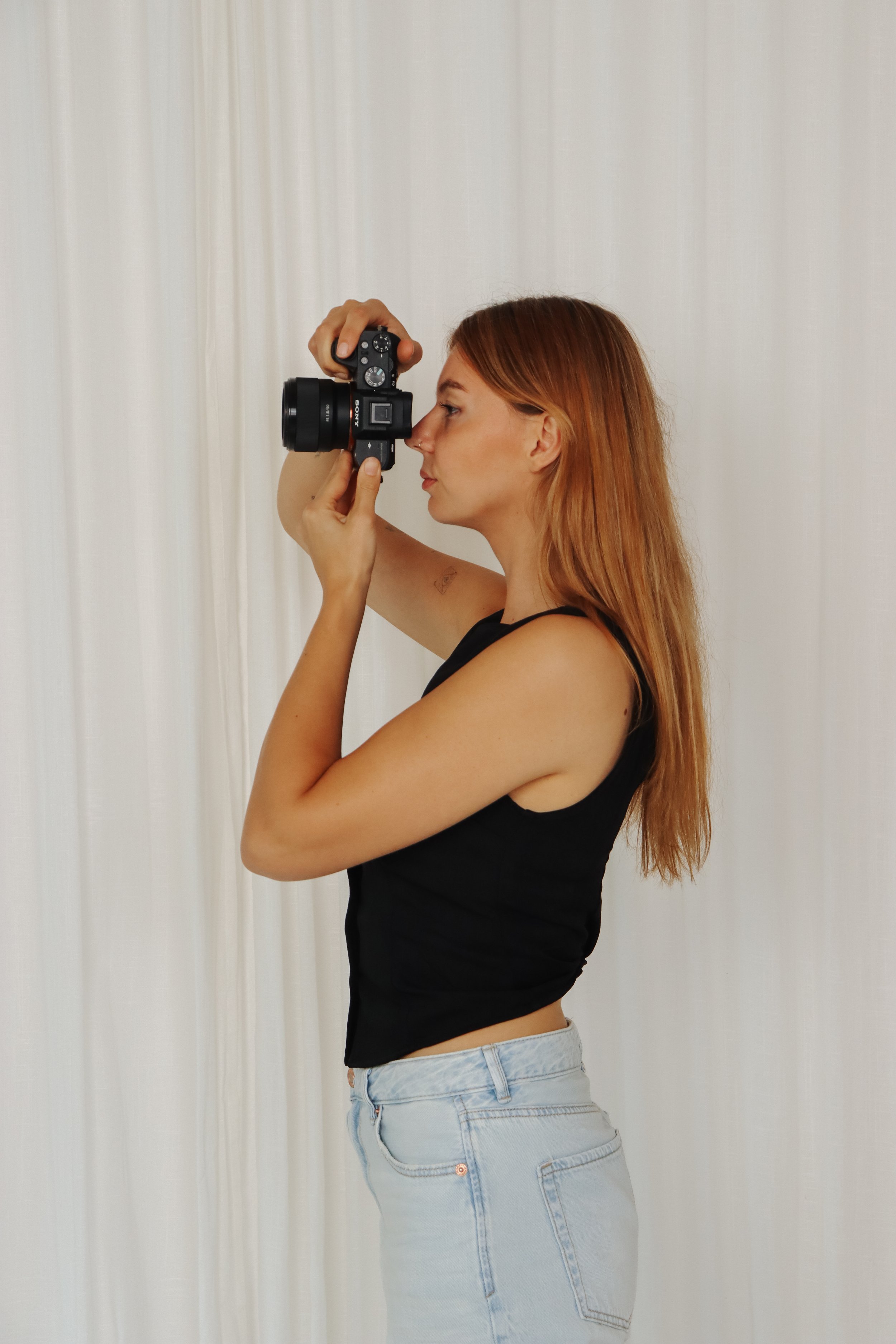 Vrouw met rood haar die een camera vasthoudt en foto's maakt, staand voor een witte achtergrond.