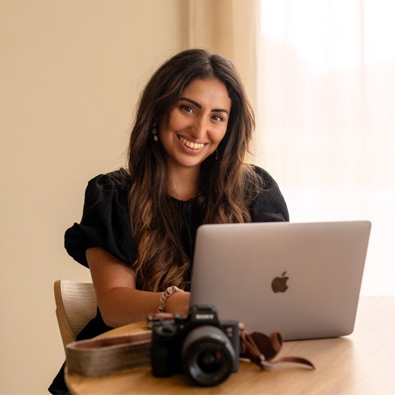 Vrouw zit aan tafel met laptop en camera, lachend naar de camera. Branding shoot