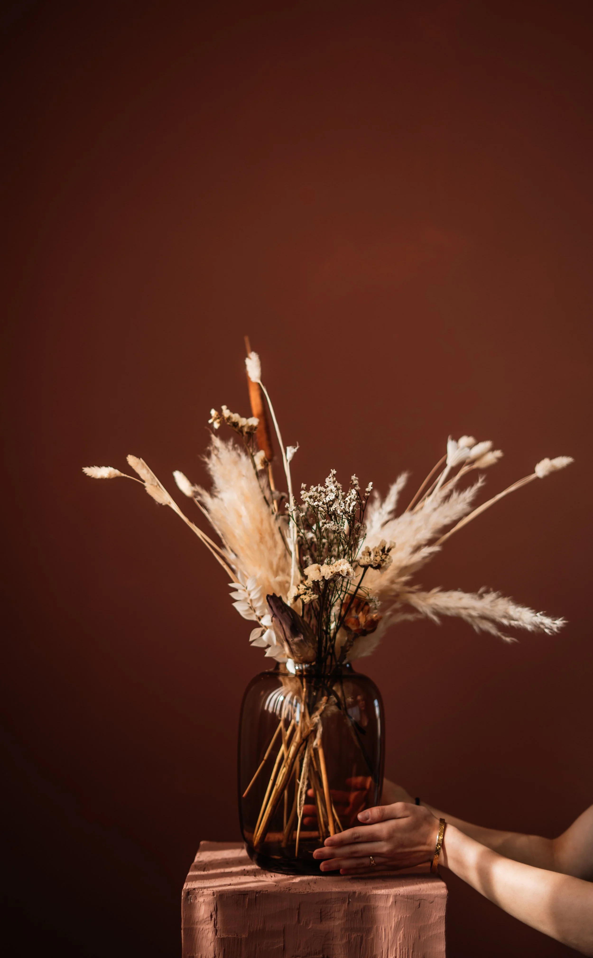 Vrouw die een donkerbruine glazen vaas met gedroogde bloemen vasthoudt op een houten oppervlak, met een effen bruine achtergrond. Floweris
