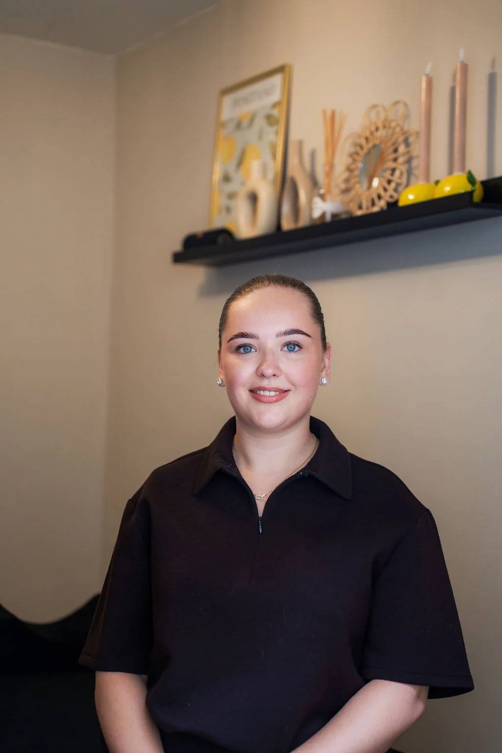 Een vrouw met een zwarte blouse staat binnen voor een beige muur met een zwart plankje met decoraties. Beautysalon.