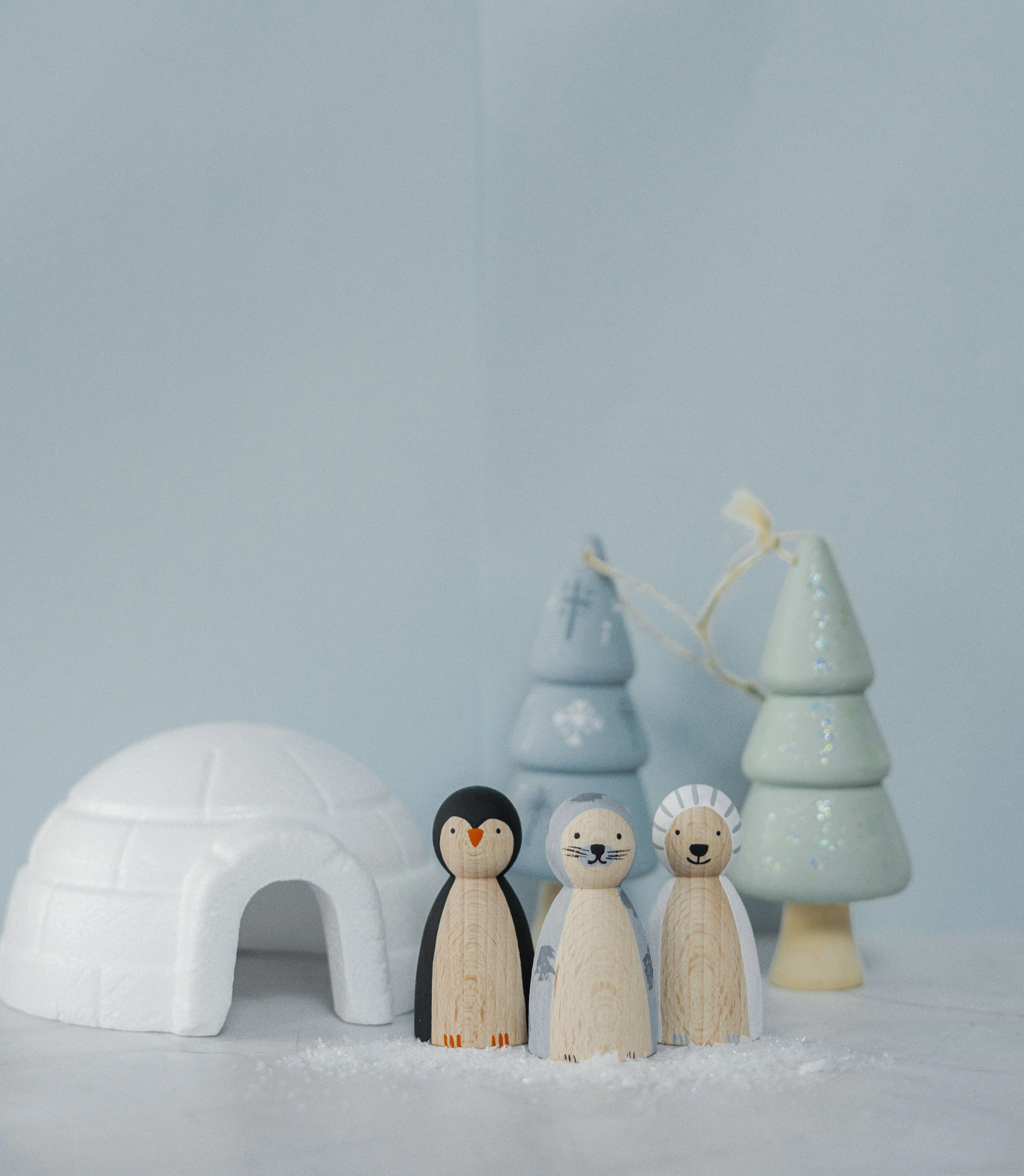 Decoratieve kersthuis, drie houten kerstfiguren van een pinguïn, ijsbeer en leeuw, en twee kerstbomen in pastelkleuren versierd met glitter, op een witte ondergrond.