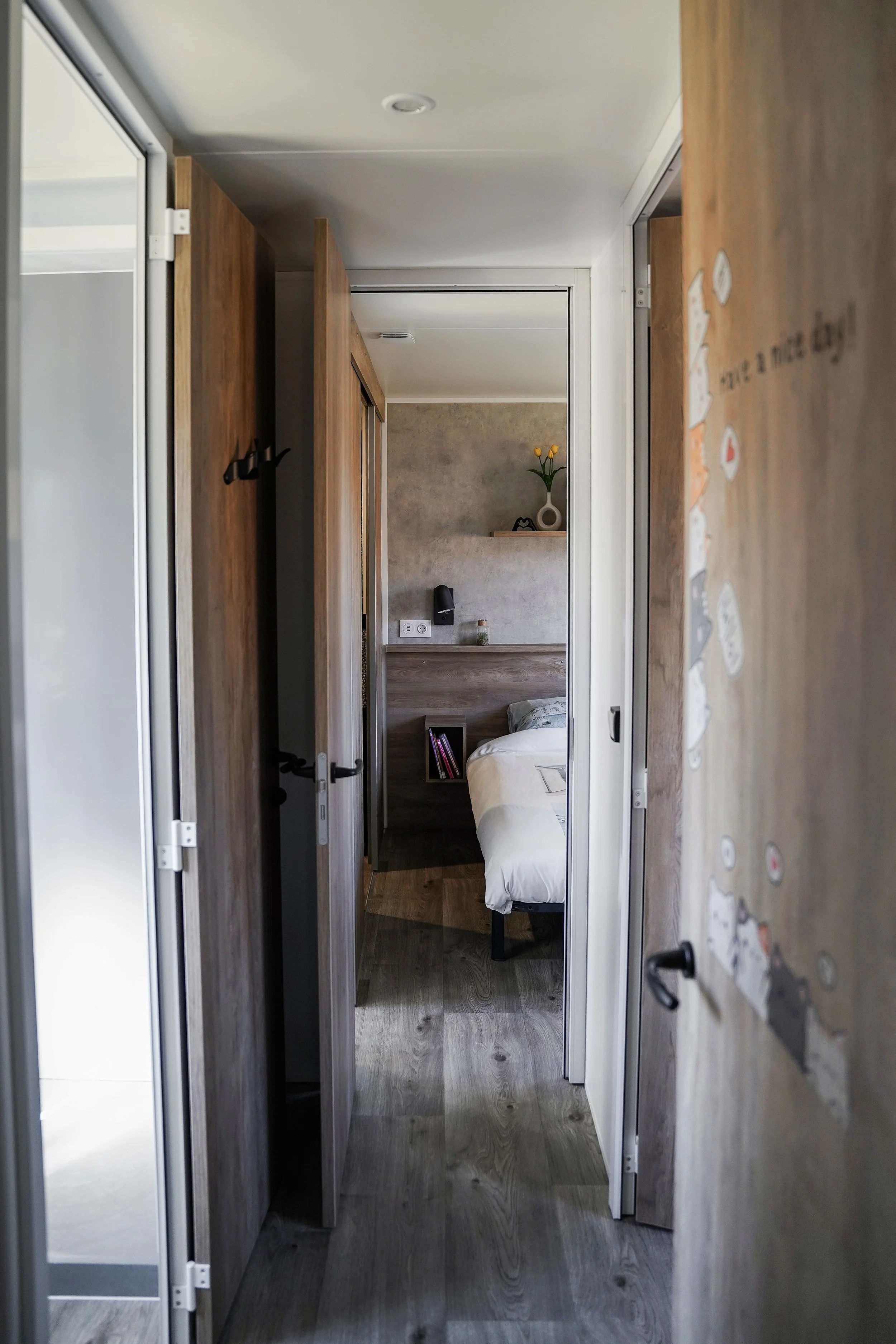 Een inkijkje van een gang richting een slaapkamer met een wit bed, een nachtkastje, en decoraties, inclusief een vaas met gele bloemen. Vakantiepark. Chalet.