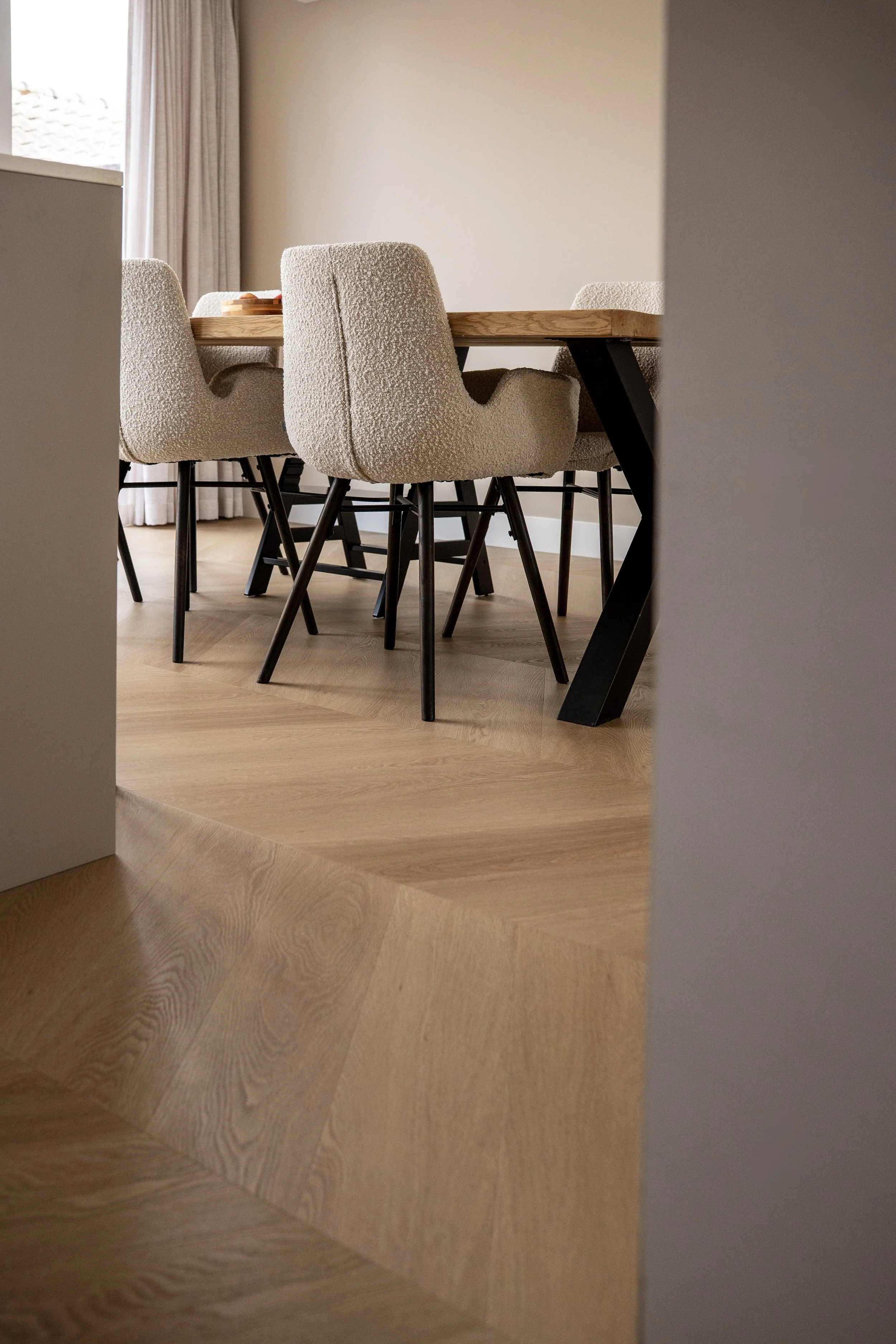 Moderne eettafel met vijf beige beklede stoelen met zwarte poten, gelegen op een lichthouten vloer, gedeeltelijk zichtbaar door een deuropening.