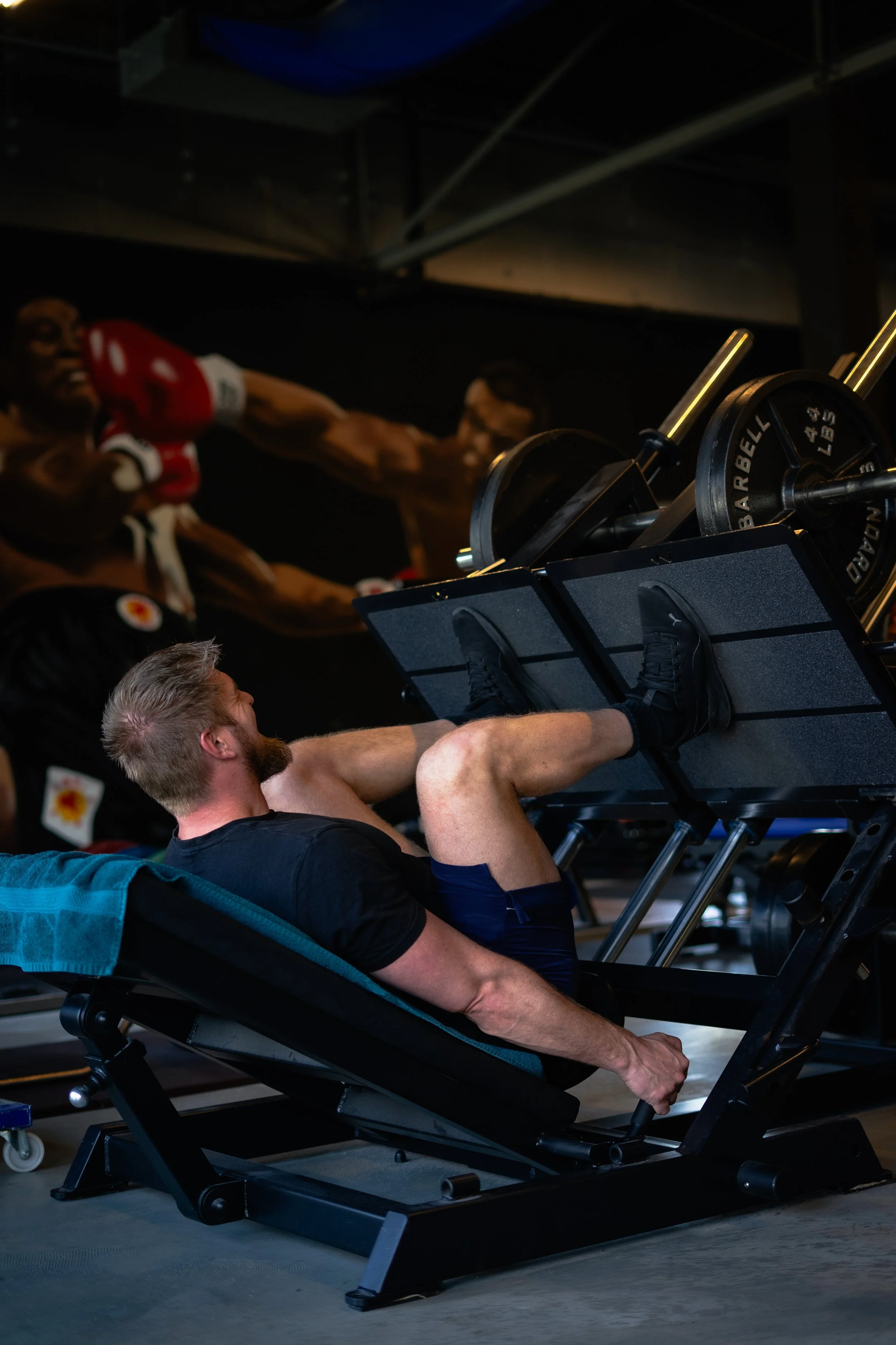 Man gebruikt een voetmachine in een sportschool terwijl een grote poster met boxers in actie op de achtergrond hangt. Sportschool Emmen. Krachttraining.