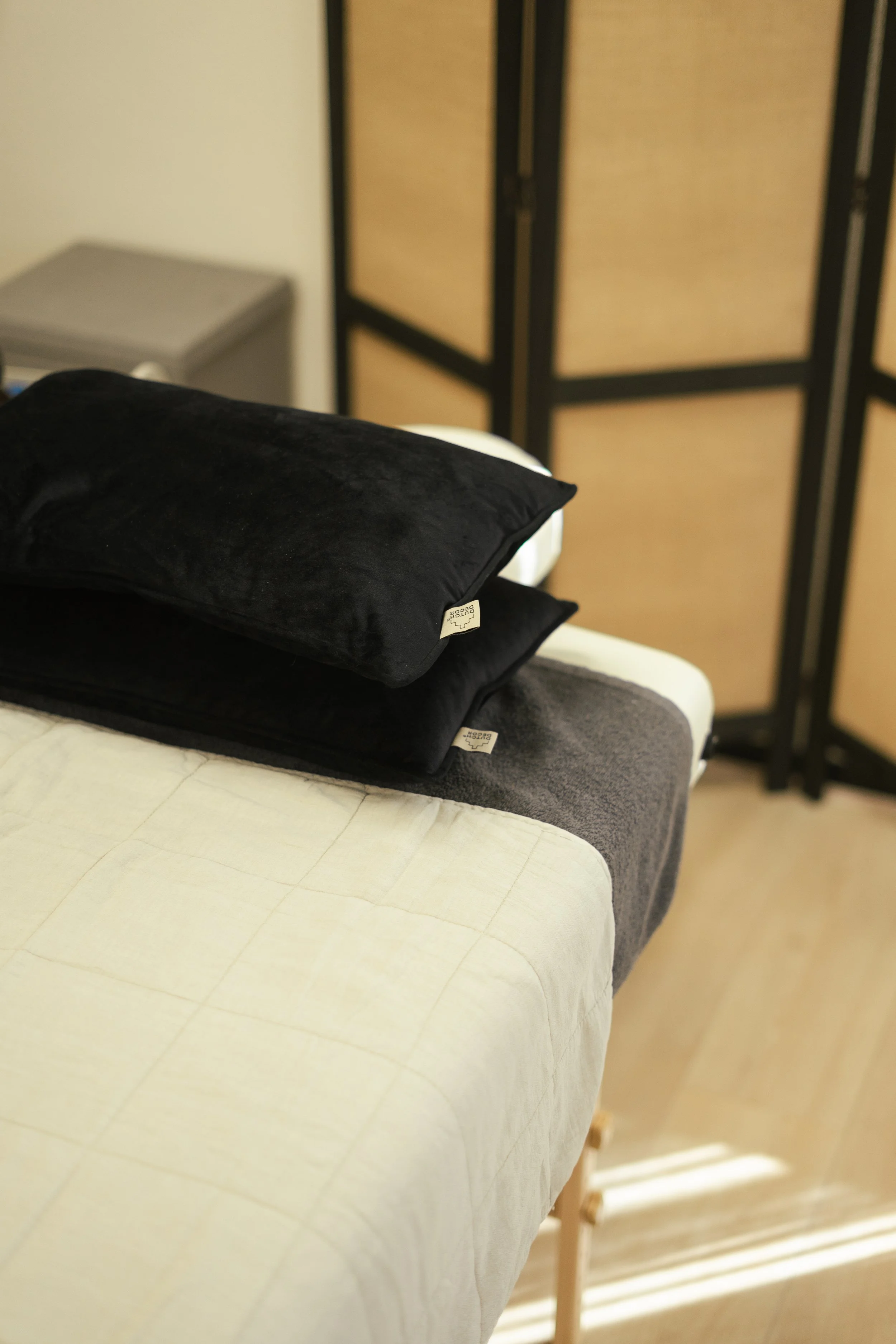 Een bed met een beige dekbedovertrek, twee zwarte kussens, en een donkere deken of laken. In de achtergrond een kamerscherm en een kleine kast.