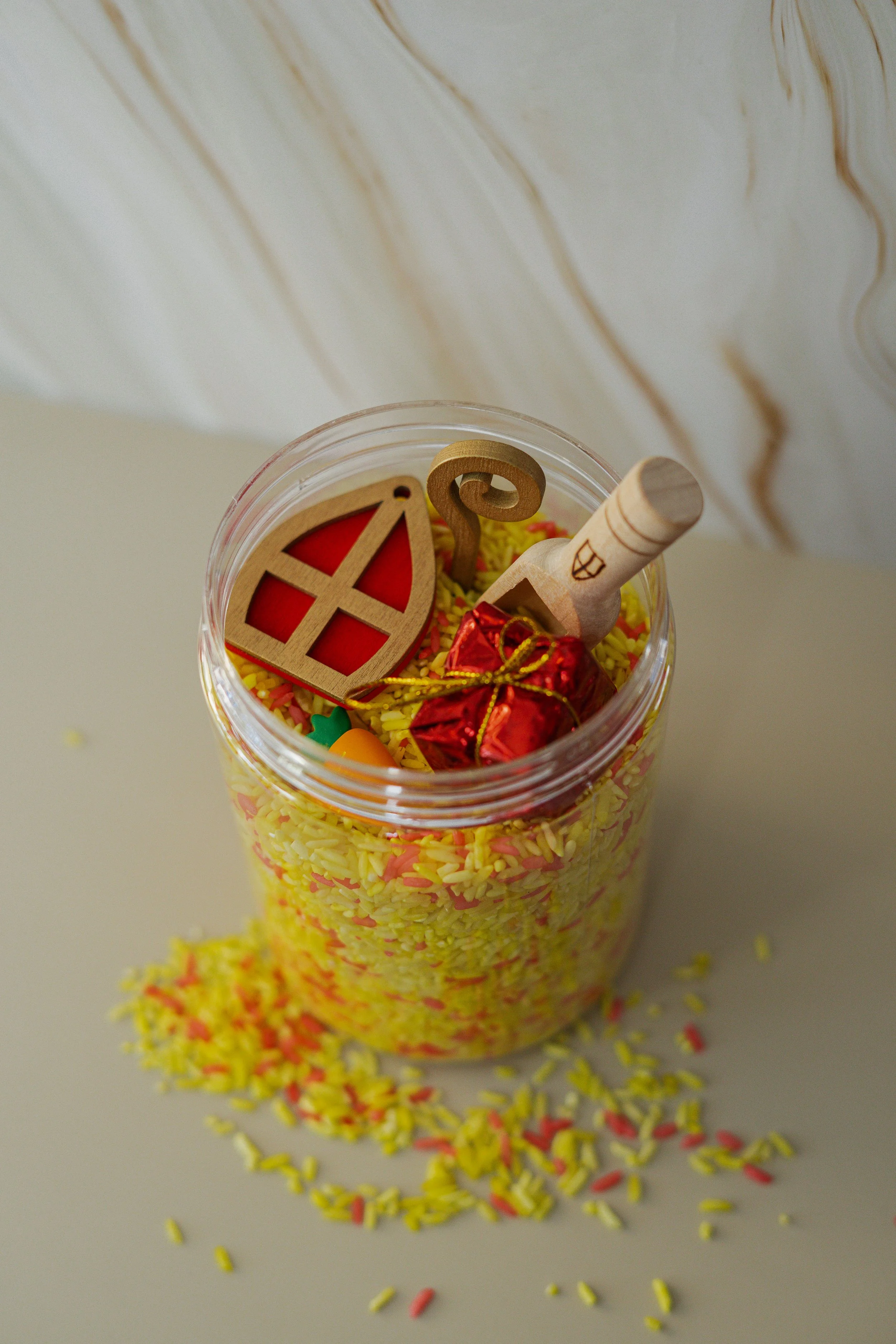 Een doorzichtige pot gevuld met gele en rode snoepjes met enkele snoepjes en mini cadeautjes eromheen.