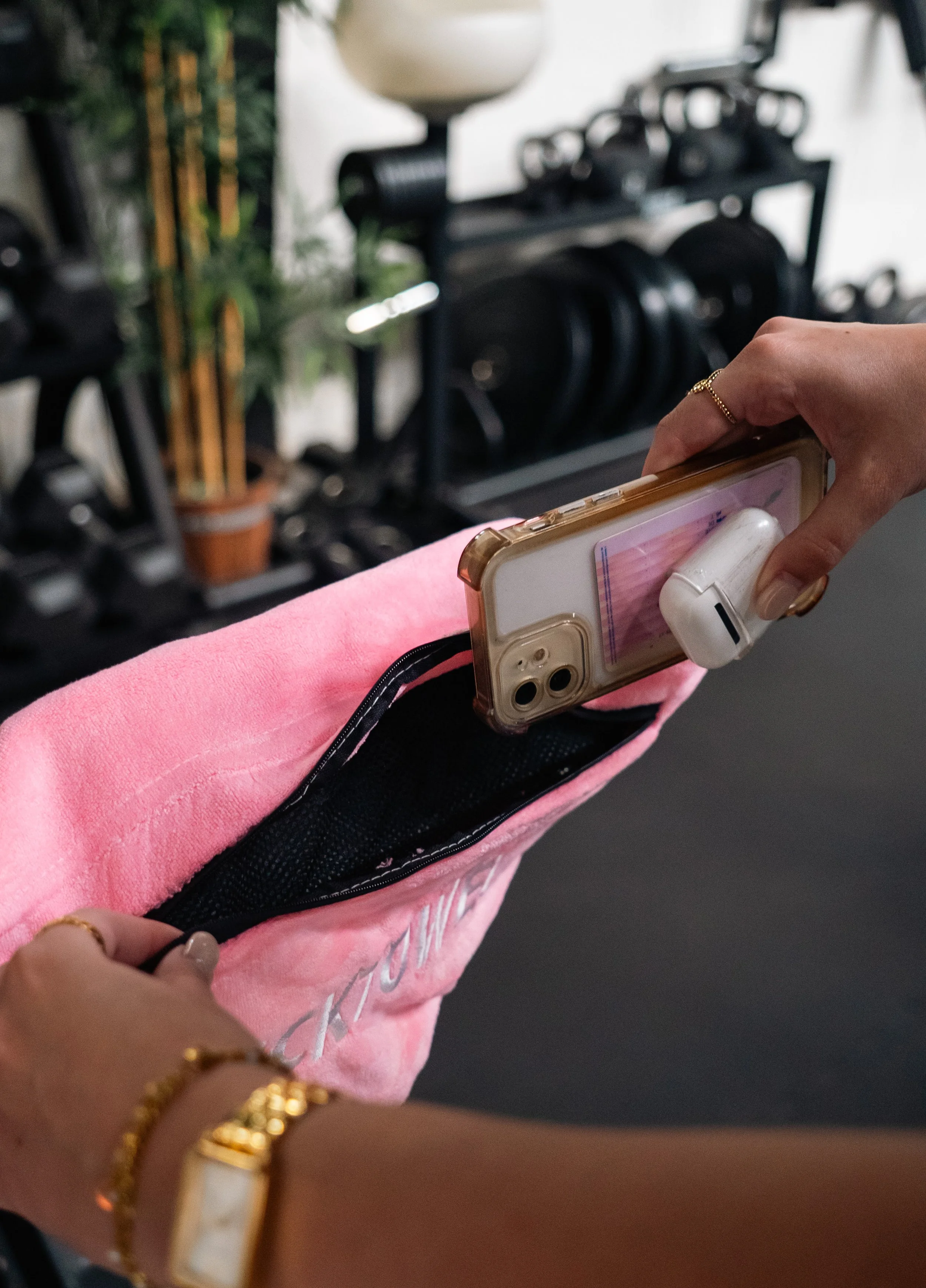 Persoon pakt een smartphone met case uit een roze tas, met AirPods in haar oor. In de achtergrond een muur met gewichten en planten. Sporthanddoek. Locktowel.