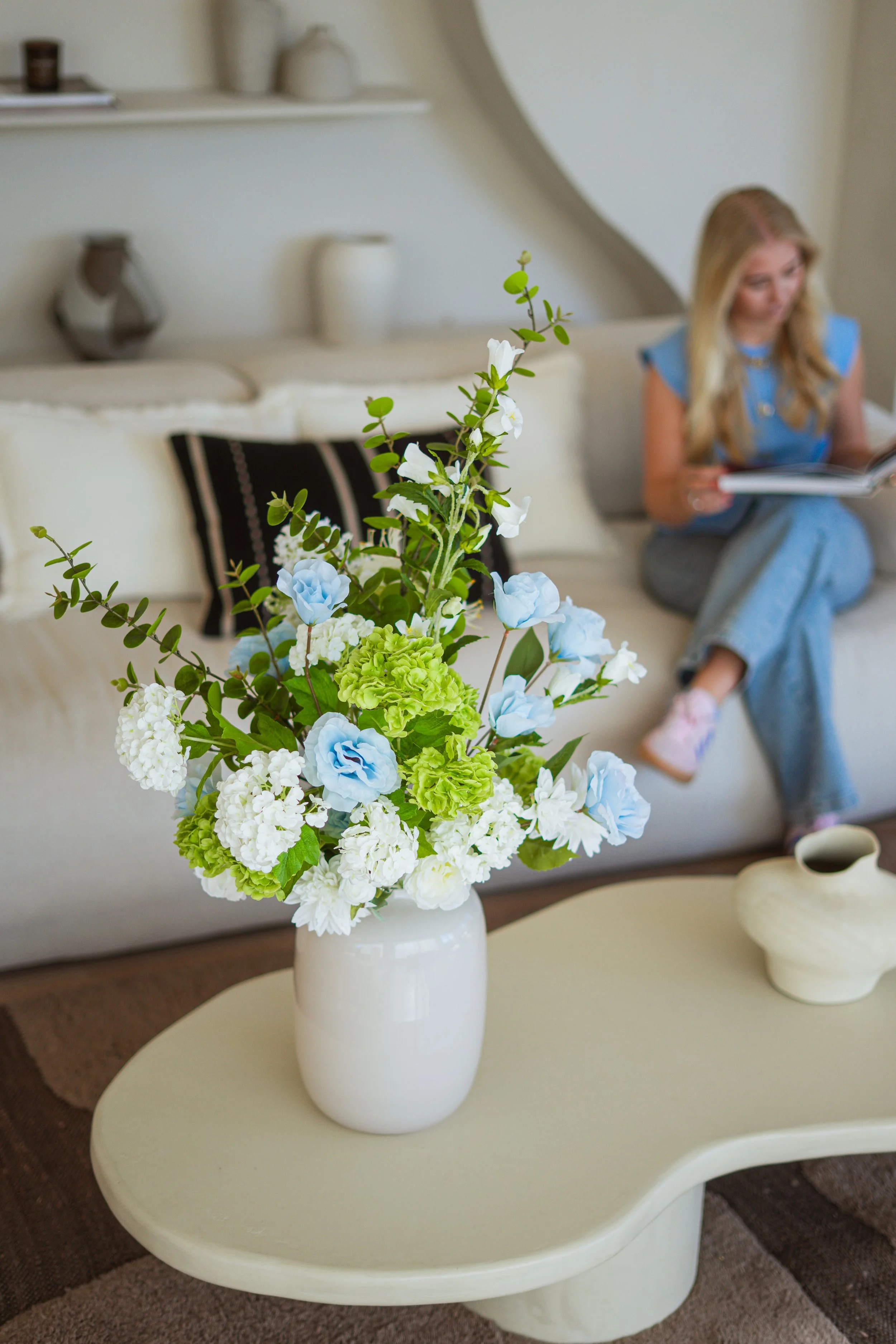 Een witte vaas met een bloemstuk van witte, blauwe en groene bloemen op een salontafel. Op de achtergrond zit een meisje op een bank die leest. Floweris