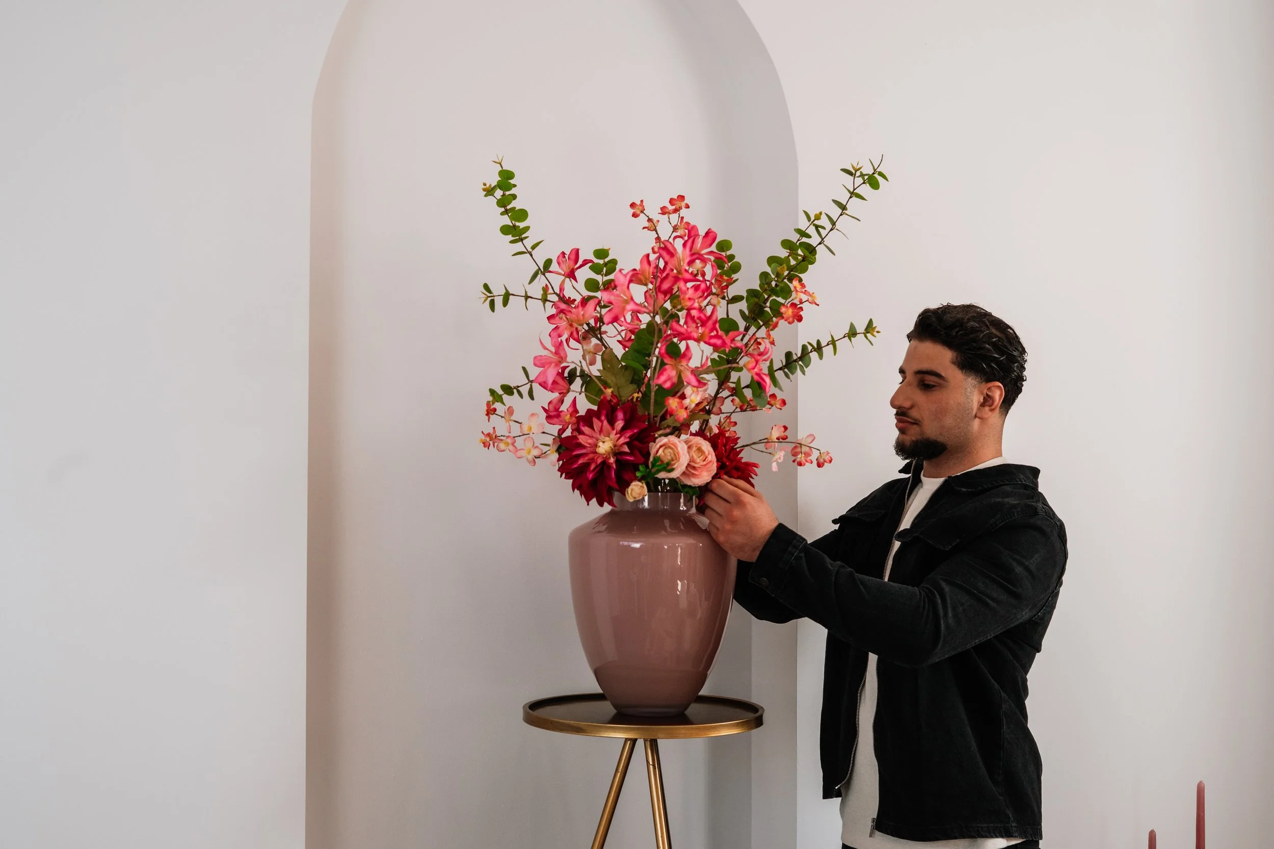 Man die een grote bloemstukken vasthoudt, bestaande uit roze en rode bloemen met groene bladeren in een grote roze vaas op een goudkleurige  Floweris