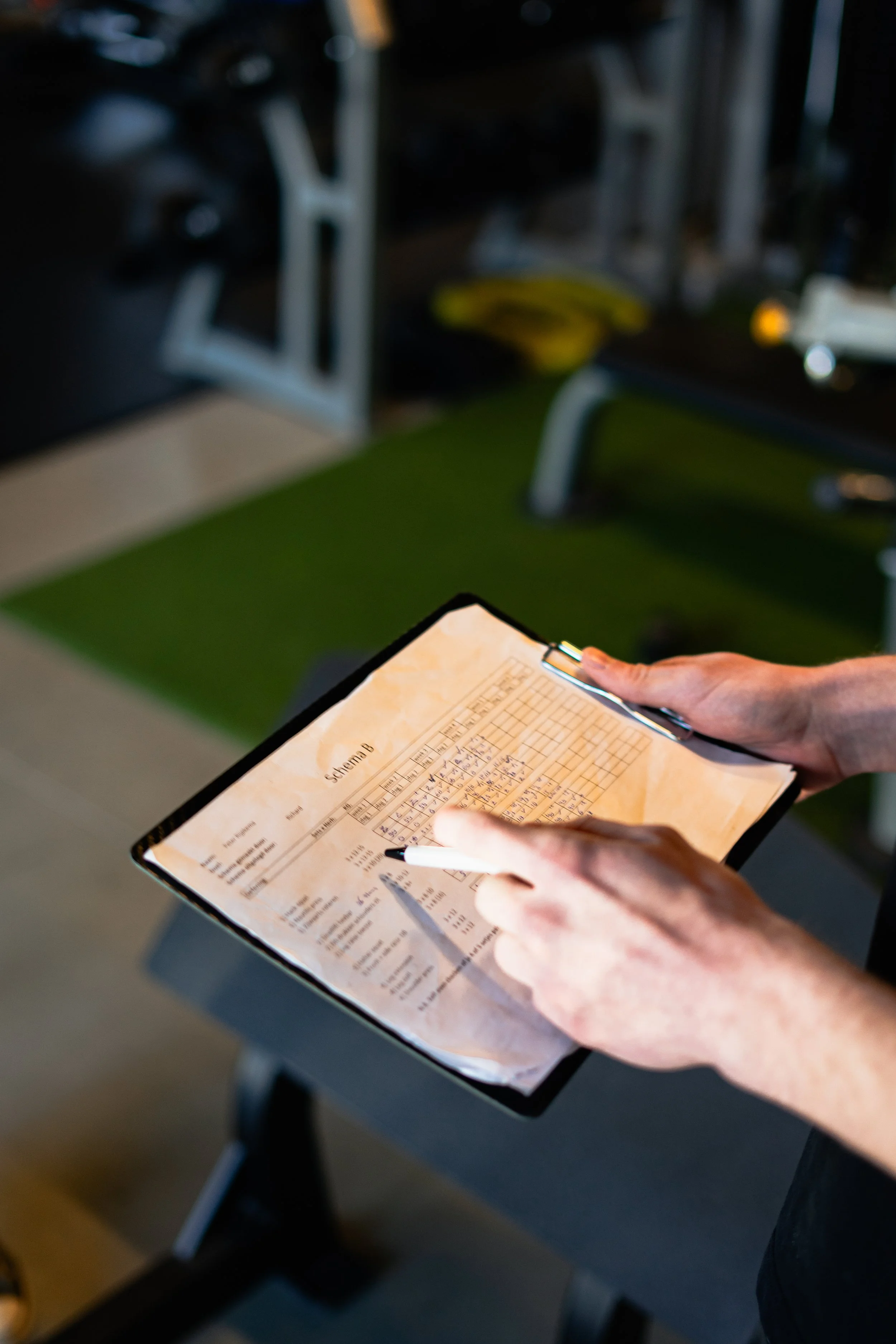 Persoon houdt een clipboard met een documenten met grafieken en tabellen, gebruikt een pen om aantekeningen te maken, in een sportschool of fitnessruimte. Sportschema